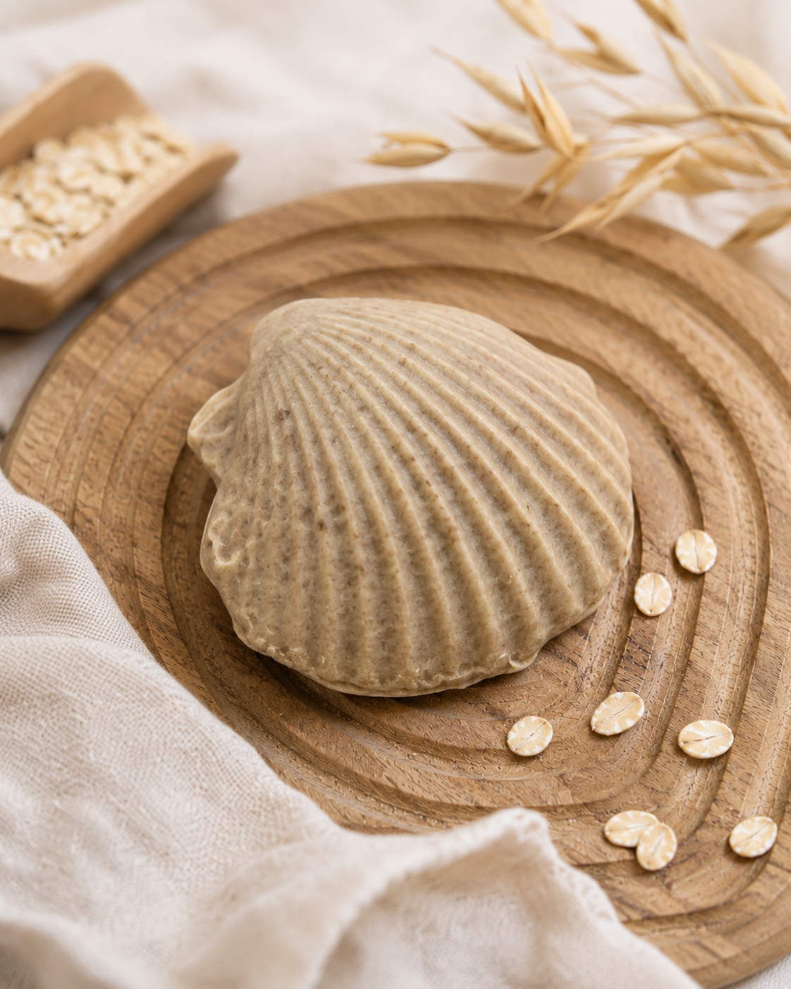 A beige, shell-shaped soap bar sits on a round wooden board, styled with scattered oats, a small wooden scoop of oats, and a soft cream-colored cloth.