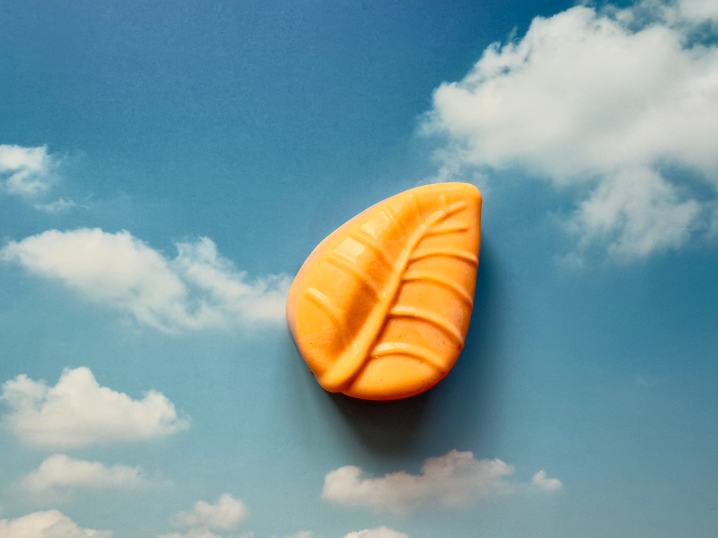 Bright orange leaf-shaped soap with subtle natural specks, photographed against a blue sky backdrop with soft white clouds.