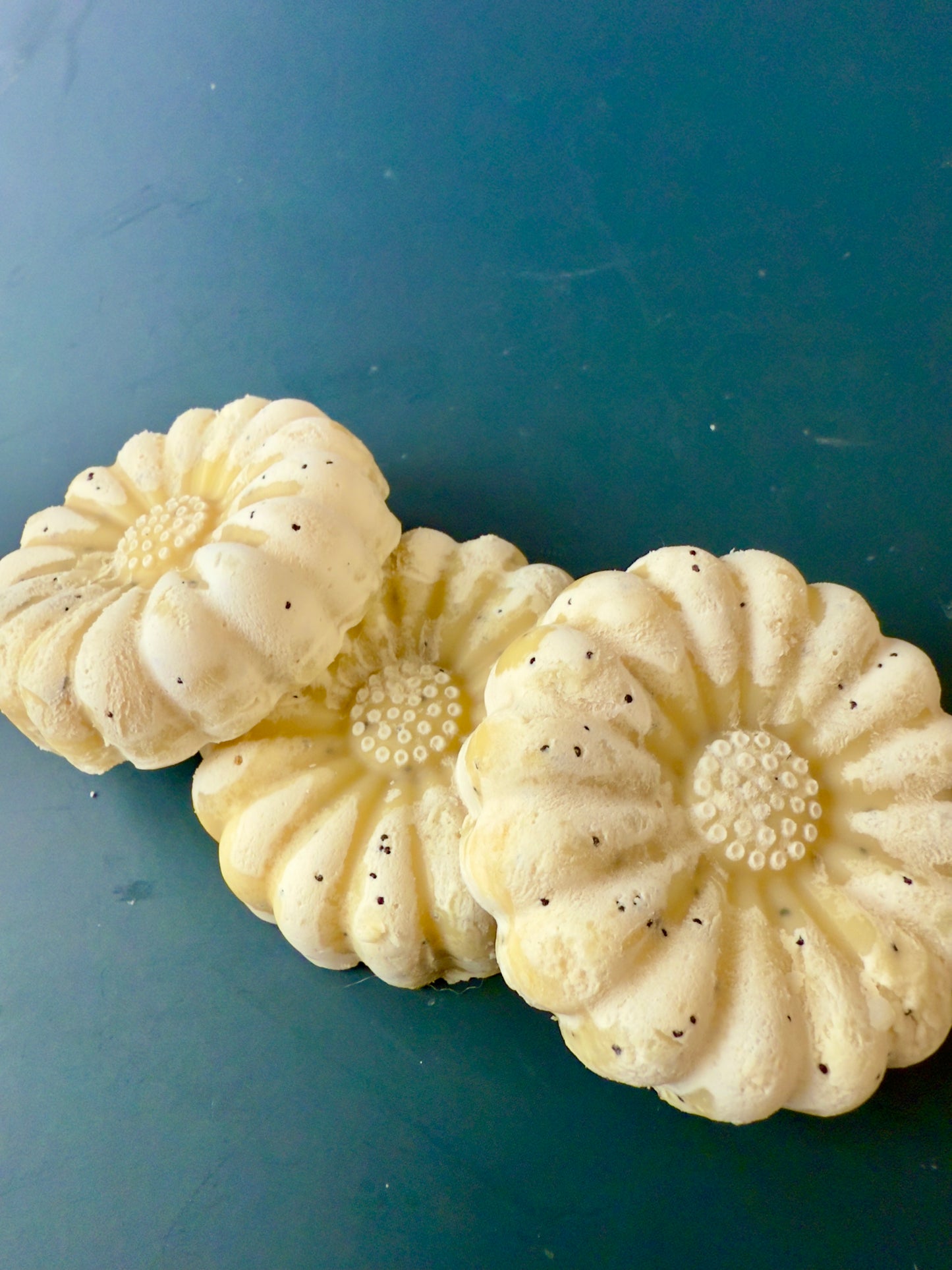 Lemon-colored and flower-shaped soaps with poppy seeds, displayed on a dark teal background.