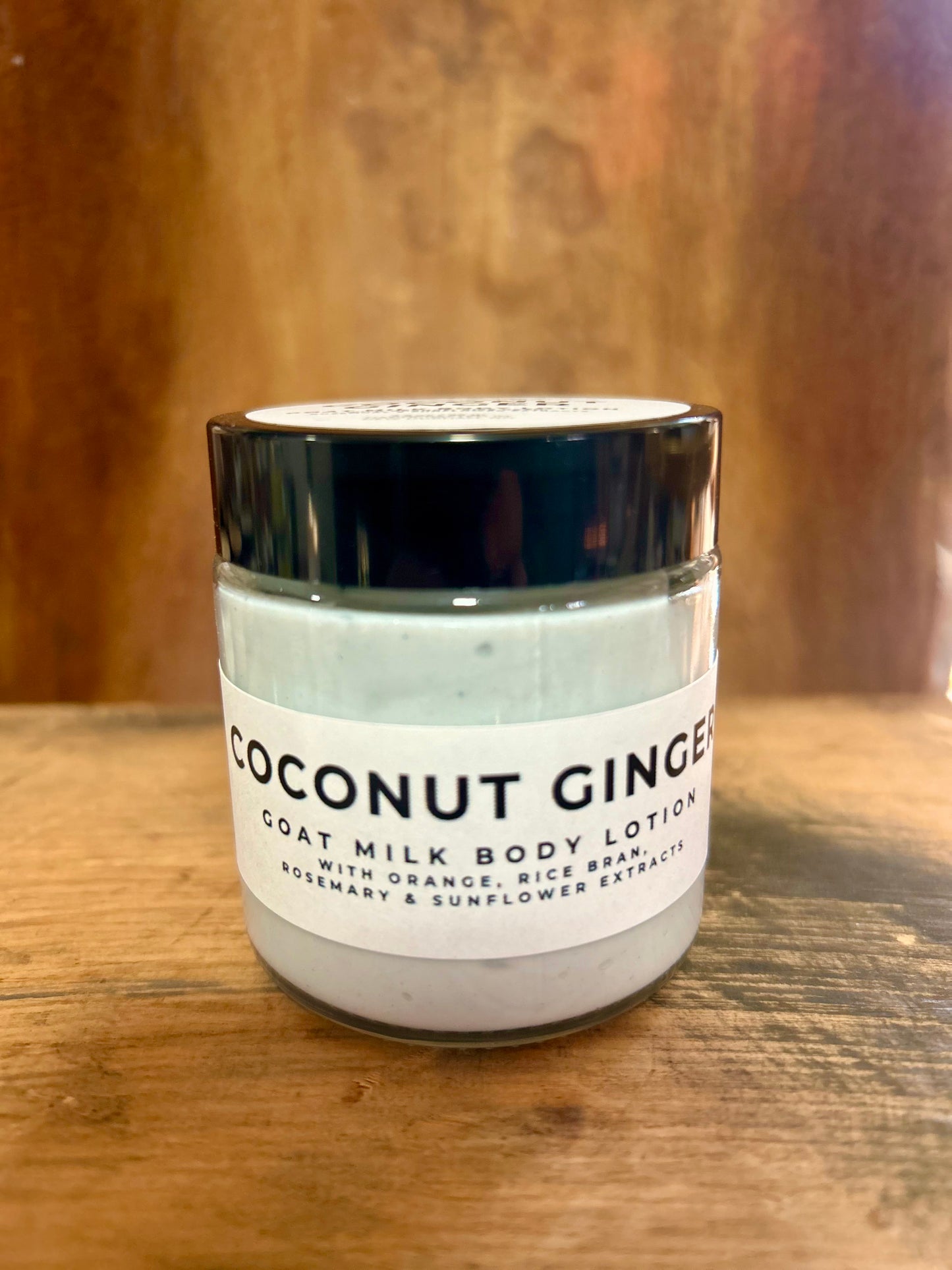 Coconut Ginger goat milk body lotion in a clear jar with a black lid and minimalist label, photographed on a warm wood background.