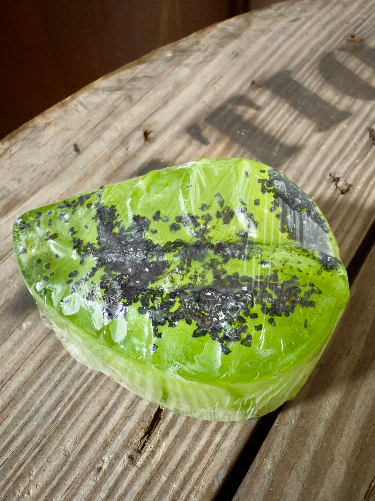 Bright green leaf-shaped soap wrapped in clear film, topped with coarse black salt, photographed on rustic wooden boards.