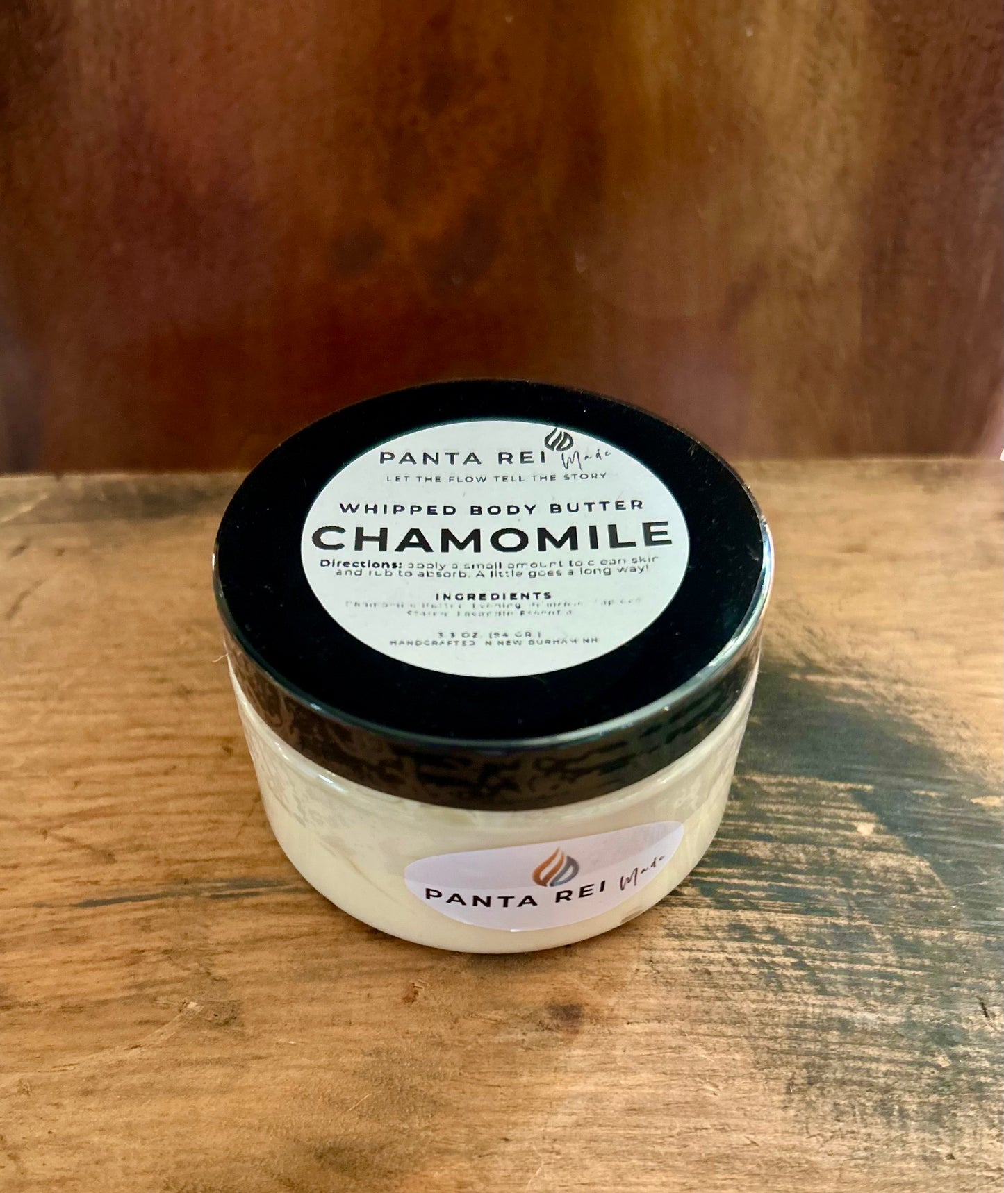 Chamomile Whipped Body Butter in a clear jar with a black lid and minimalist label, photographed on a warm wood surface.