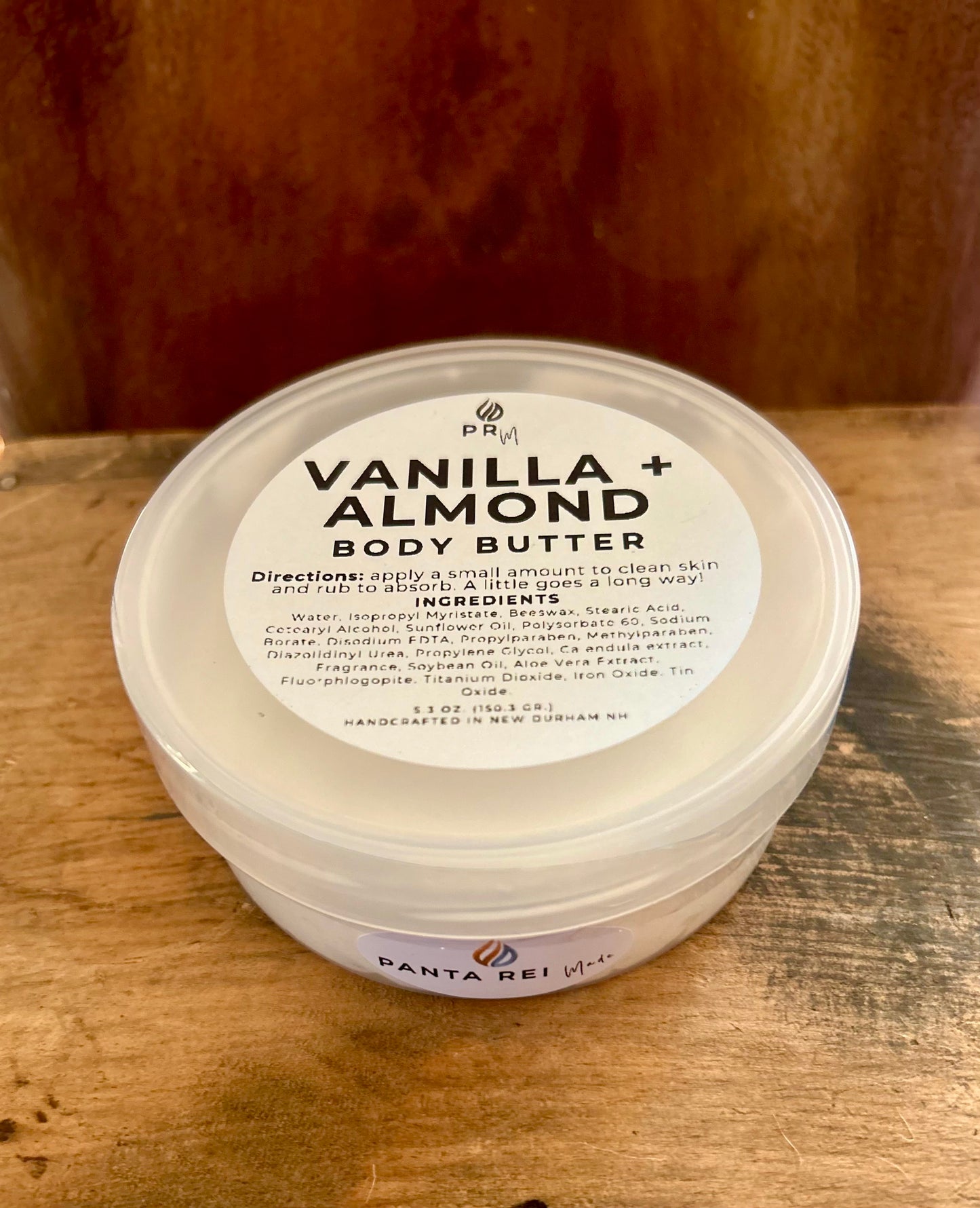 Vanilla + Almond body butter in a clear jar with a white lid and minimalist label, photographed on a warm wood background.