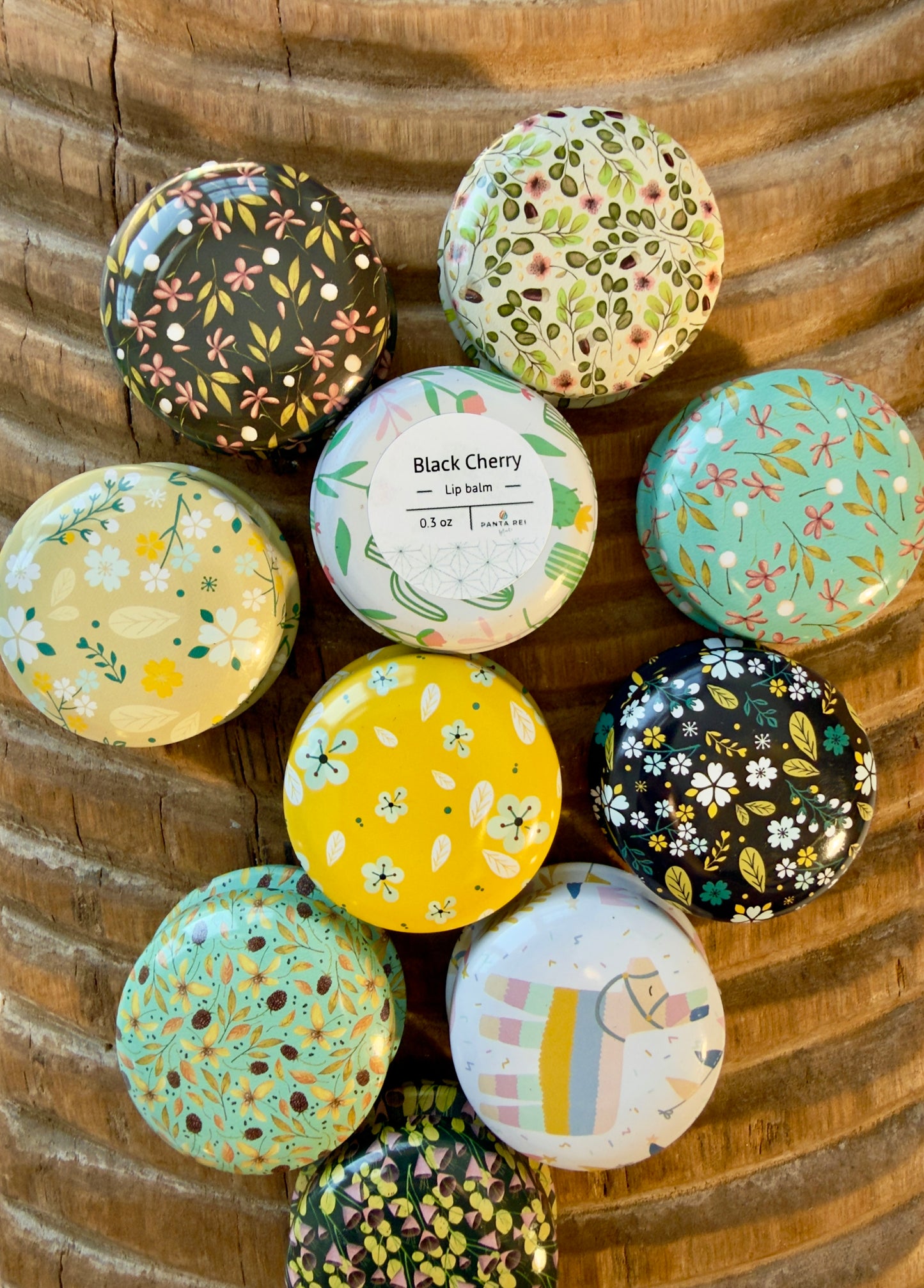 Black Cherry lip balm tin surrounded by patterned floral tins on carved wood surface.