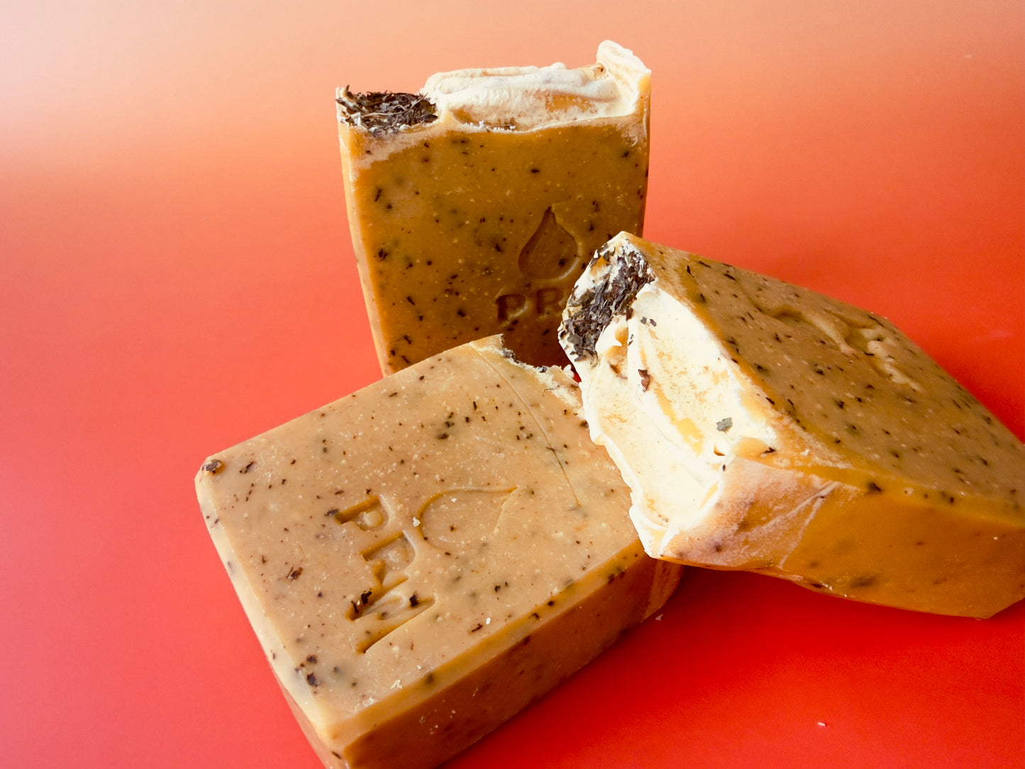 Three bars of soap with a warm golden tone, visible spearmint leaf flecks throughout, and dried spearmint leaves on top against a soft gradient background.