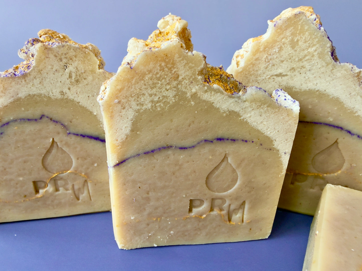 Lavender Cedarwood soap bars with PRM logo, gold and purple mica accents visible along the top, set against a blue background
