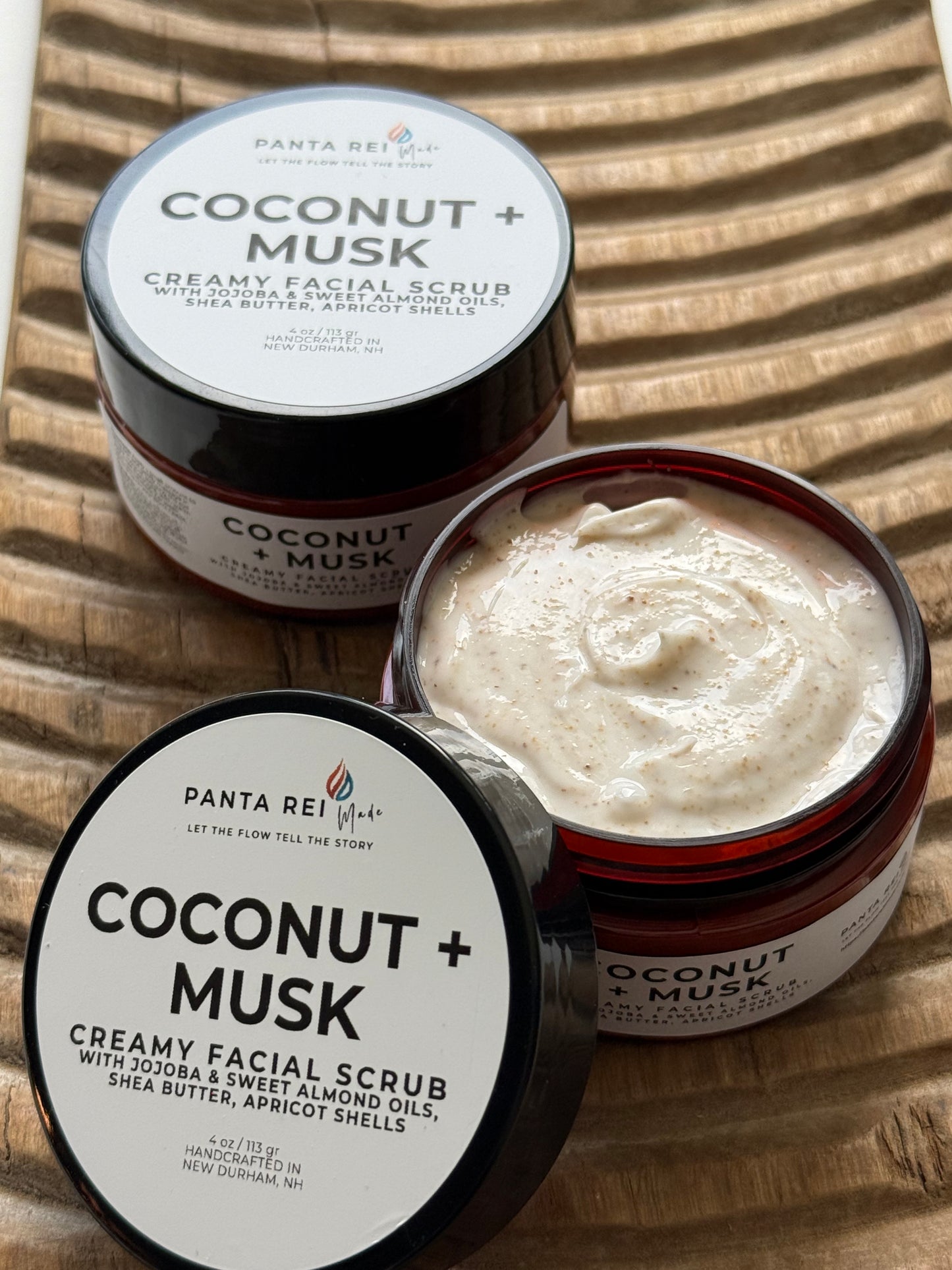 Coconut + Musk creamy facial scrub in amber jars with black lids on a wooden tray.