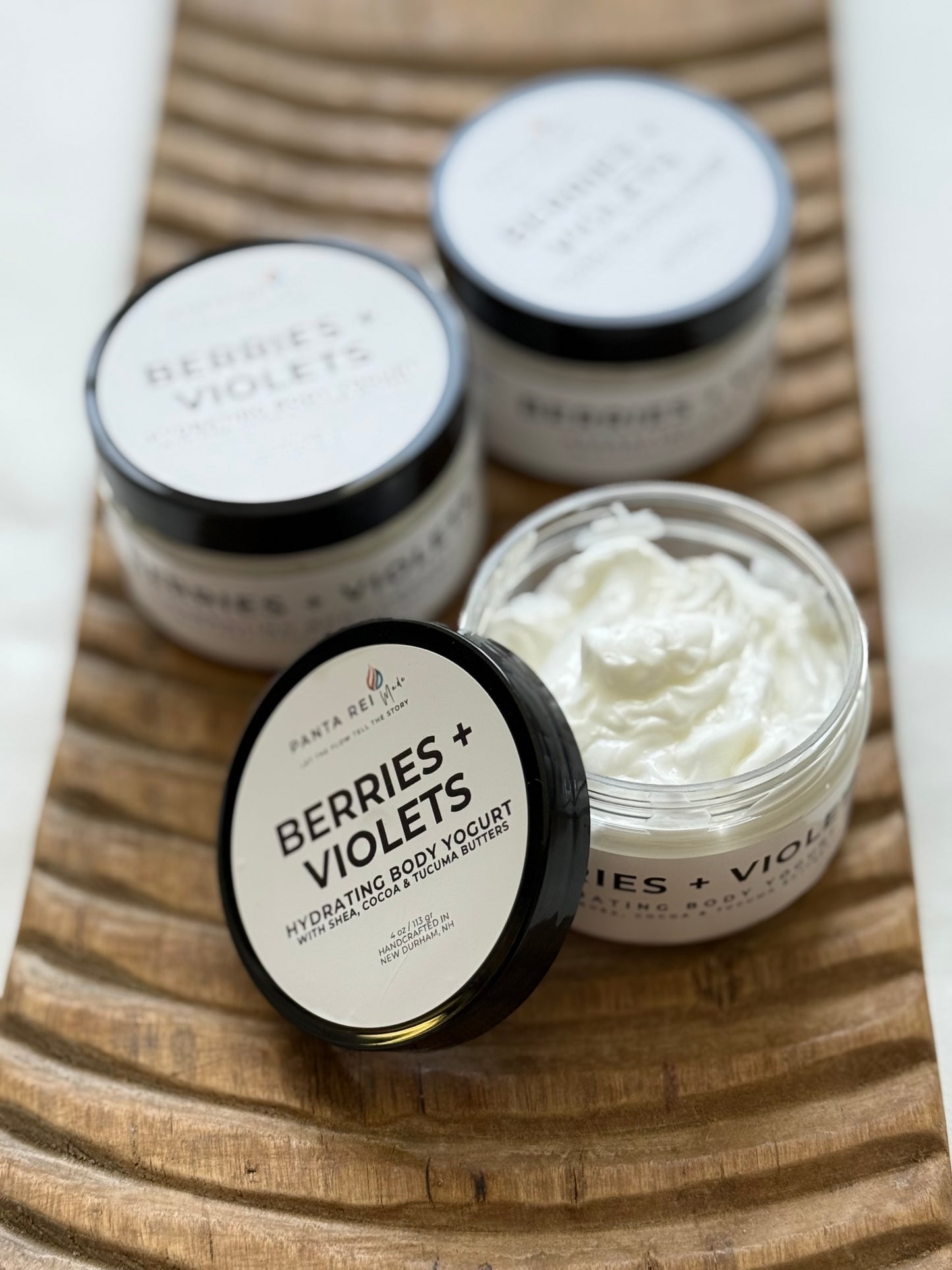 Berries + Violets hydrating body yogurt in white jars with black lids on a wooden tray.