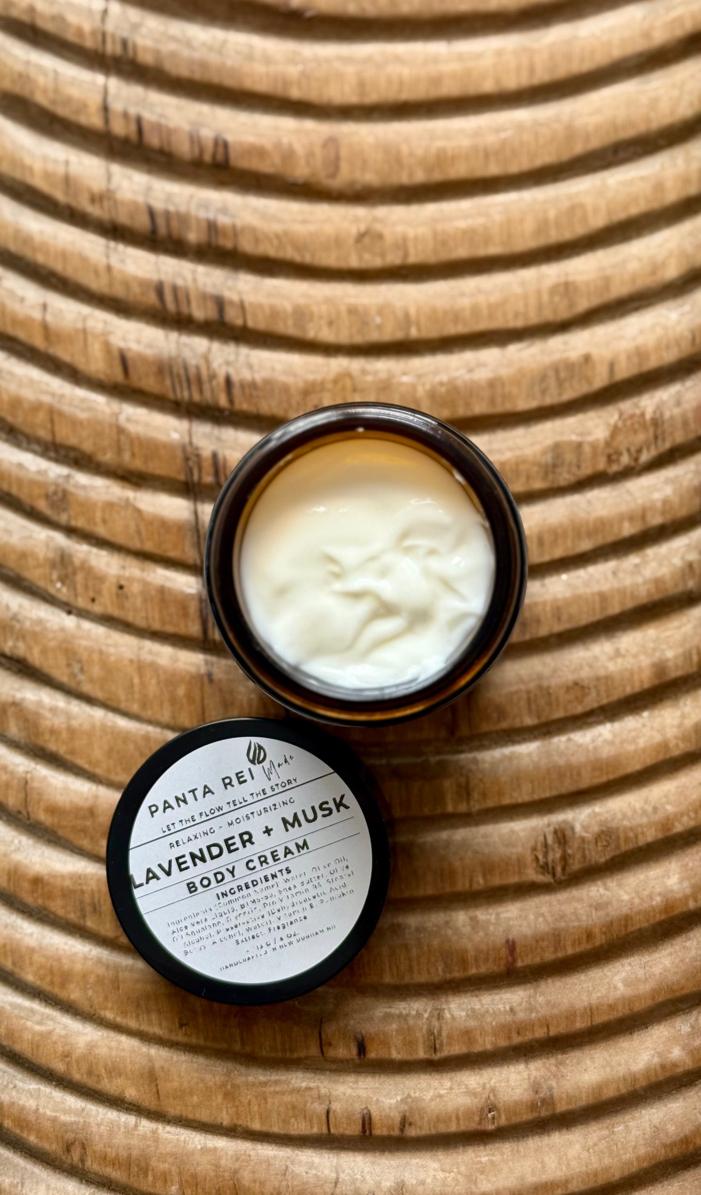 Amber-glass jar of Lavender + Musk Body Cream on carved wooden surface, shown open with smooth cream texture and matching lid.