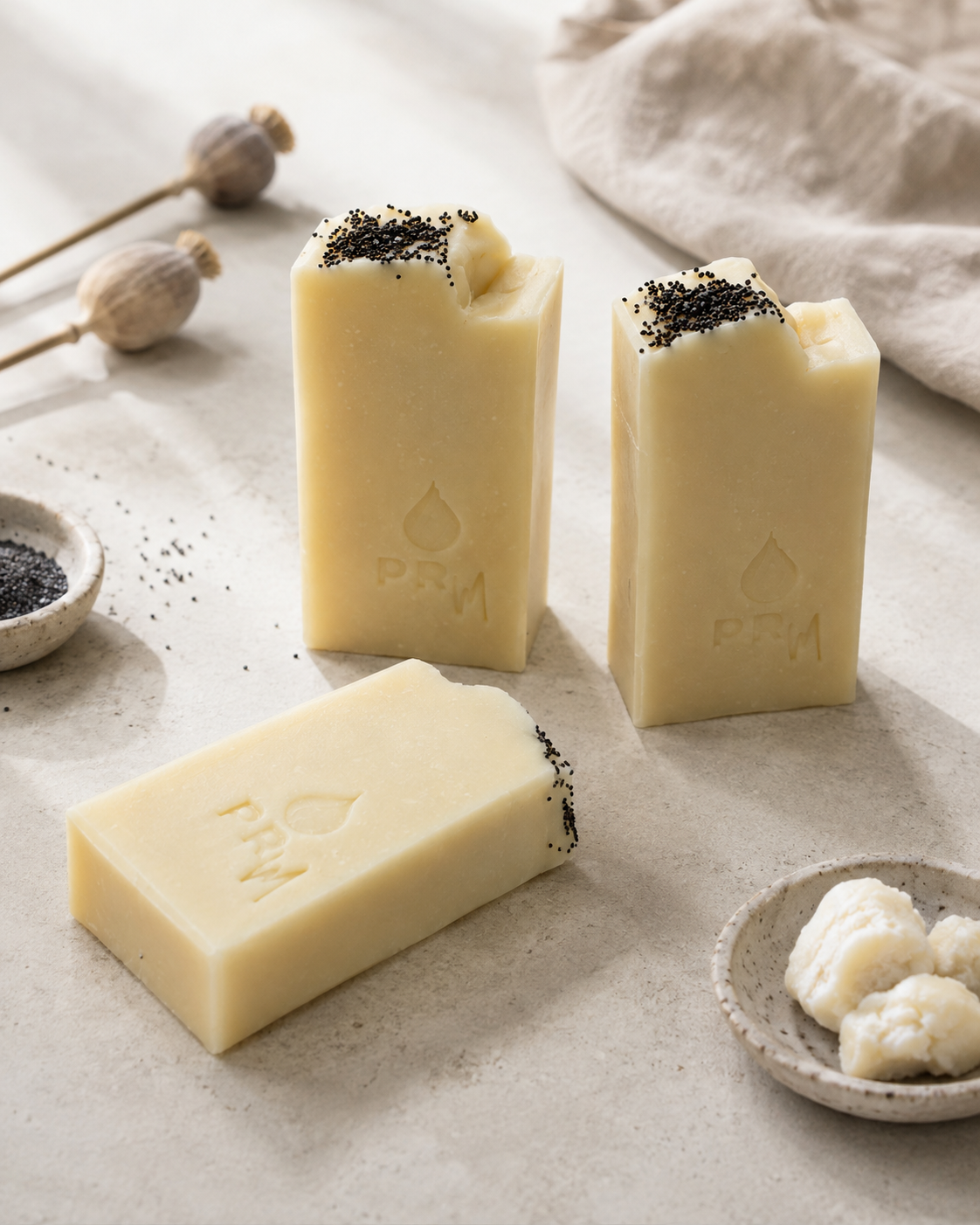 Three bars of pale yellow soap with black seed topping, branded with 'PRM', arranged on a textured beige surface with small bowls of ingredients and dried pods.