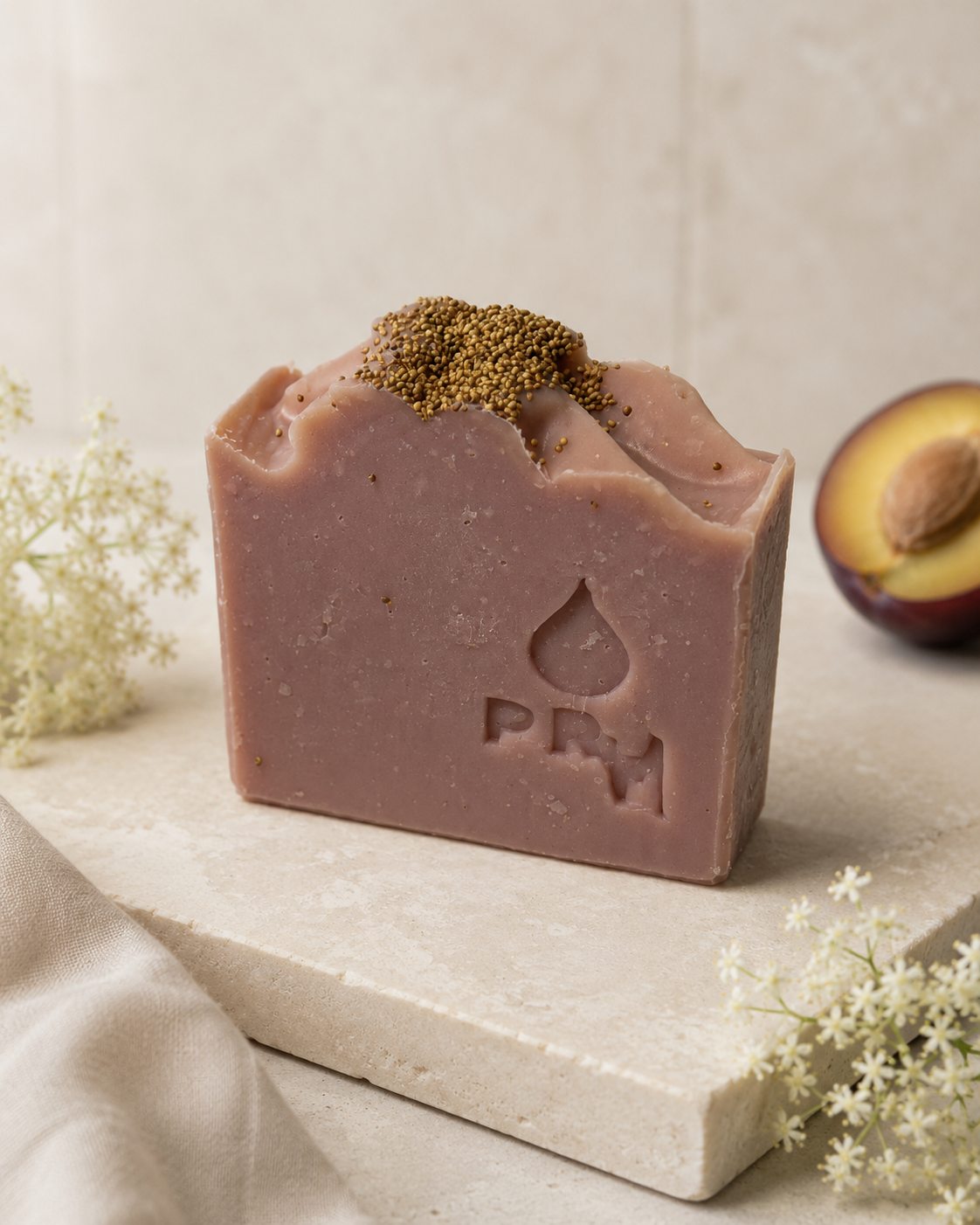 A rectangular bar of dusty pink-colored handmade soap with a textured top and a pile of brown seeds on top, resting on a light stone slab. The soap features an embossed droplet logo and the letters 'PRM'.