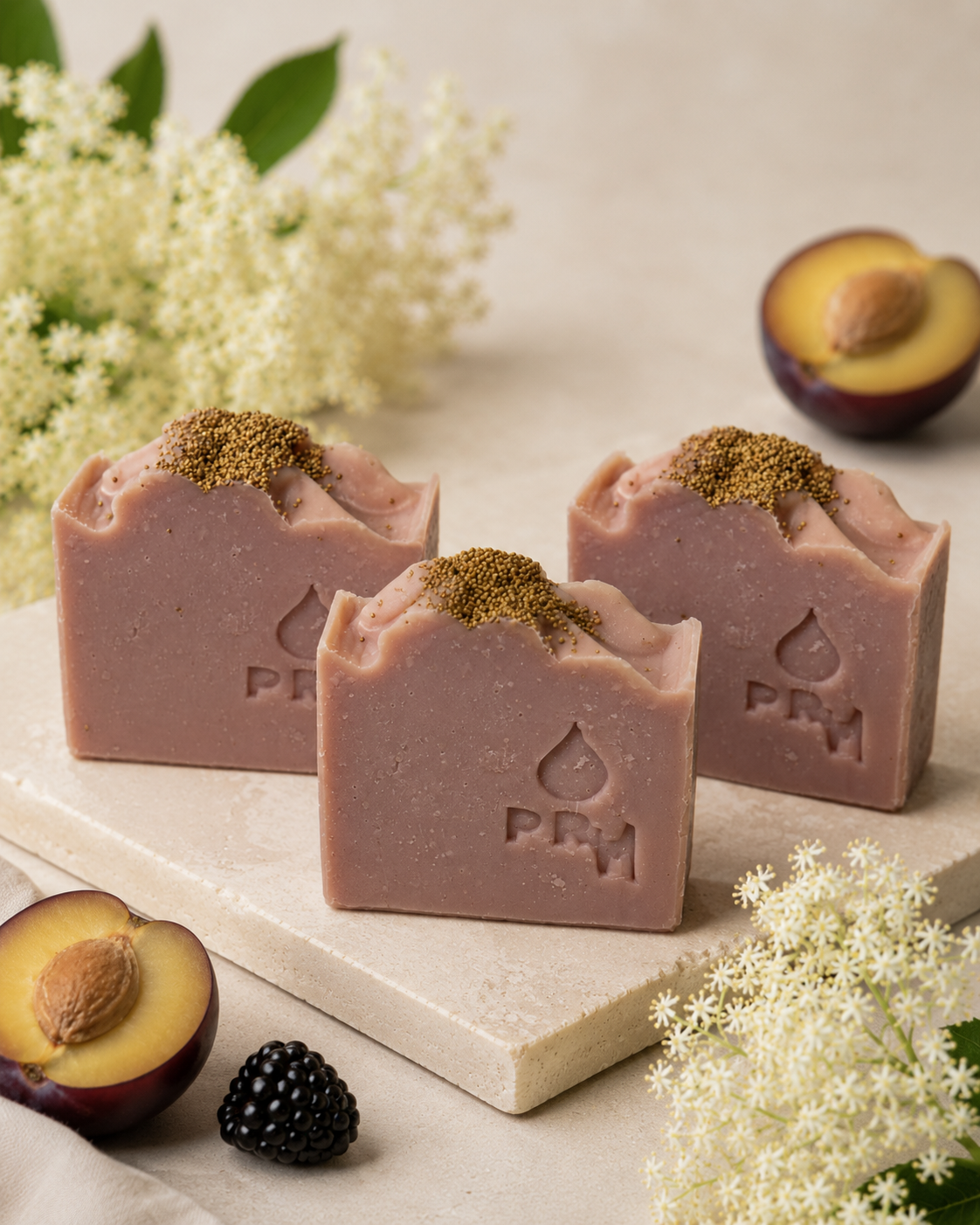Three rectangular bars of dusty pink-colored soap with a gold-brown spice dusting on top, arranged on a light stone slab with halved plums, a blackberry, and white flowers in the background.