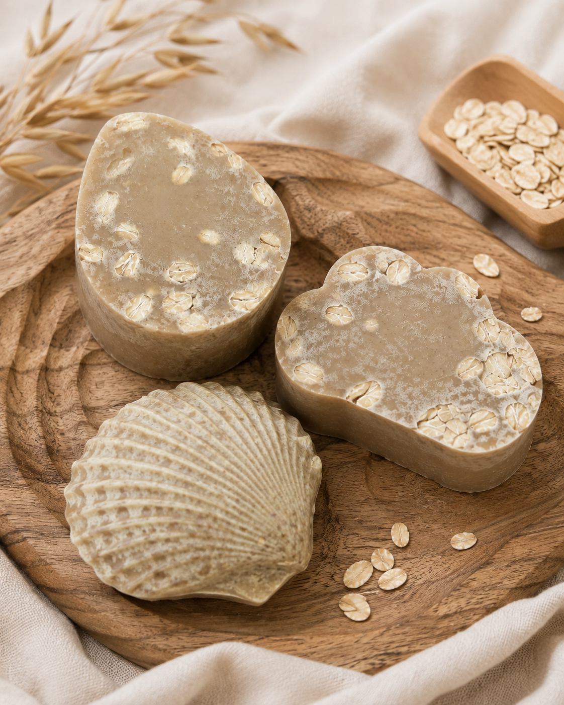 Three handmade soap bars with organic oat shapes and embedded oats, displayed on a wooden tray alongside oats in a wooden dish, and neutral fabric.