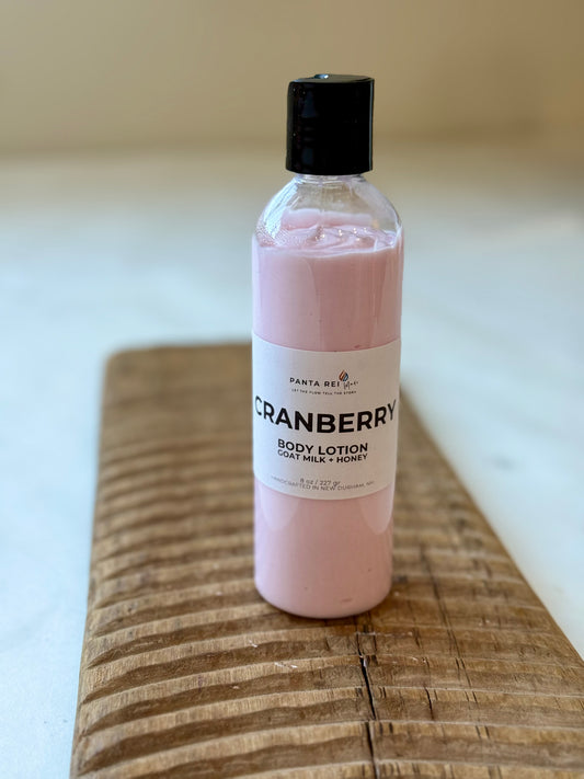 Tall clear bottle of pink Cranberry body lotion with a black cap, placed on a carved wooden board against a light background