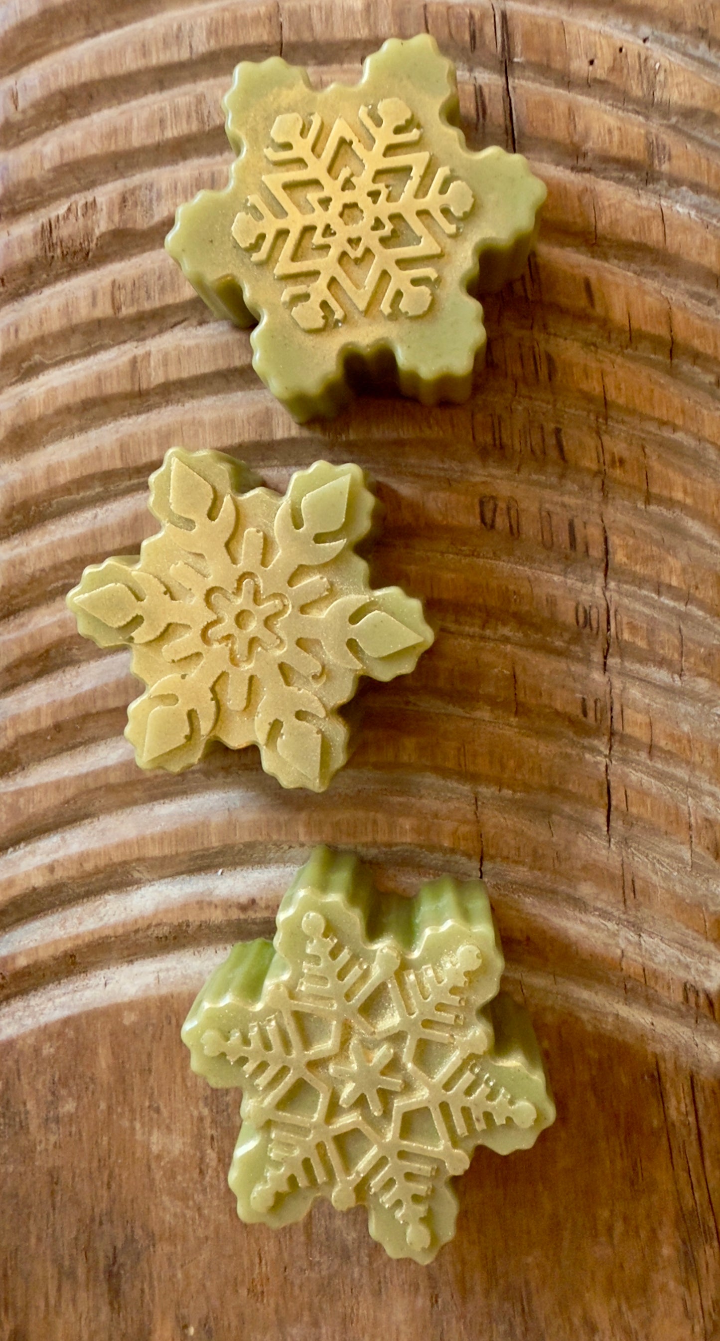 Green Christmas tree-shaped soap on a wooden surface. Green tree and snowflake holiday goat milk soaps with gold brushed tops and natural speckling.