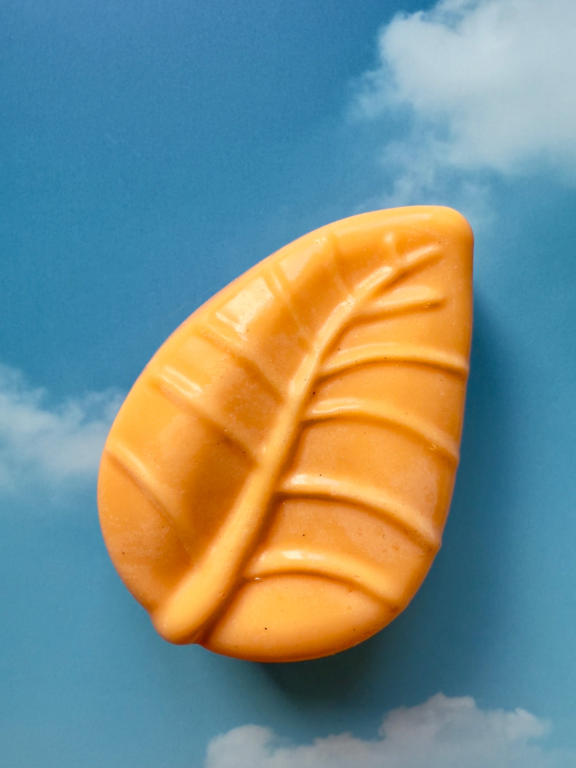 Bright orange leaf-shaped soap with subtle natural specks, photographed against a blue sky backdrop with soft white clouds.