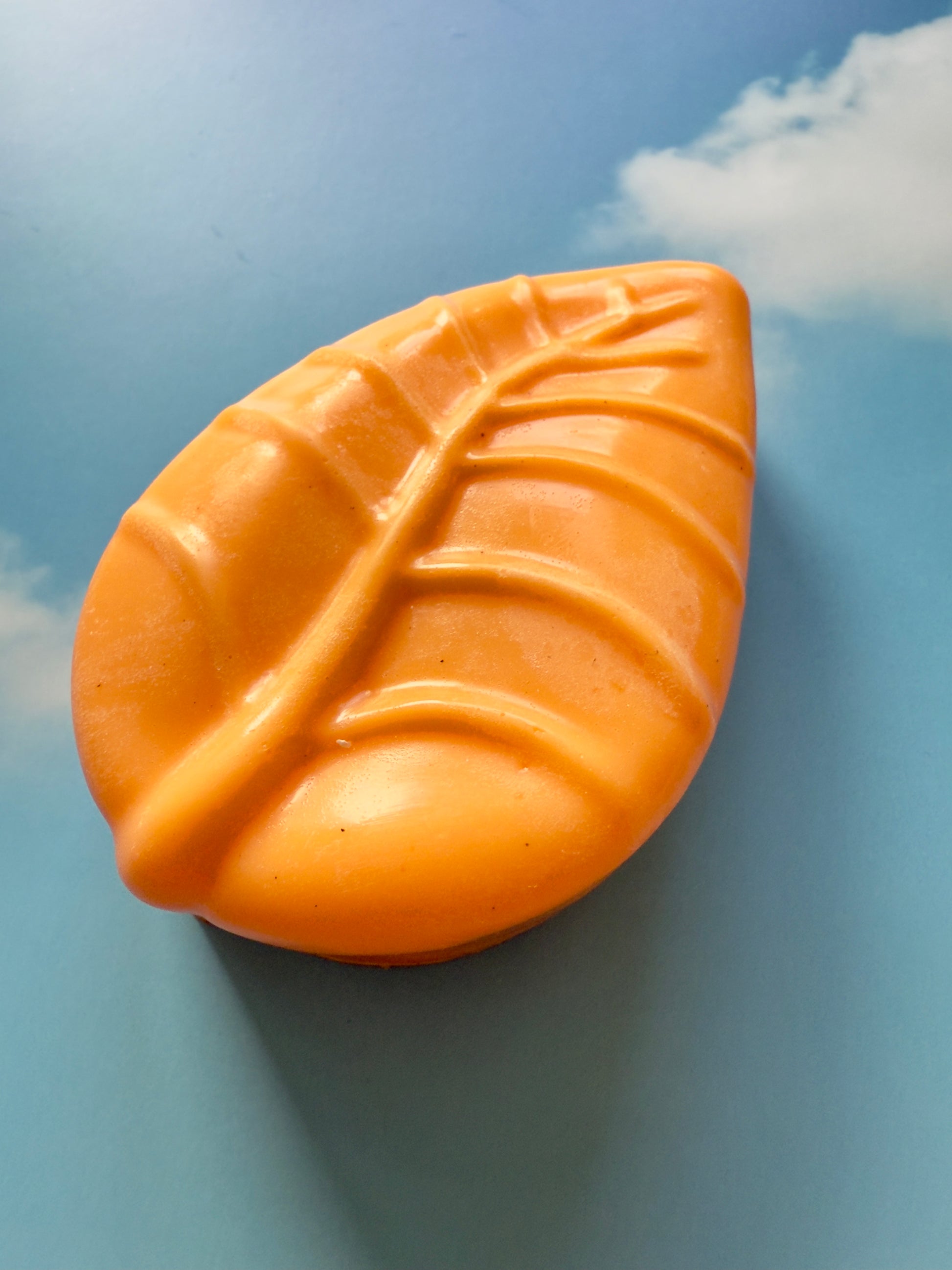 Bright orange leaf-shaped soap with subtle natural specks, photographed against a blue sky backdrop with soft white clouds.