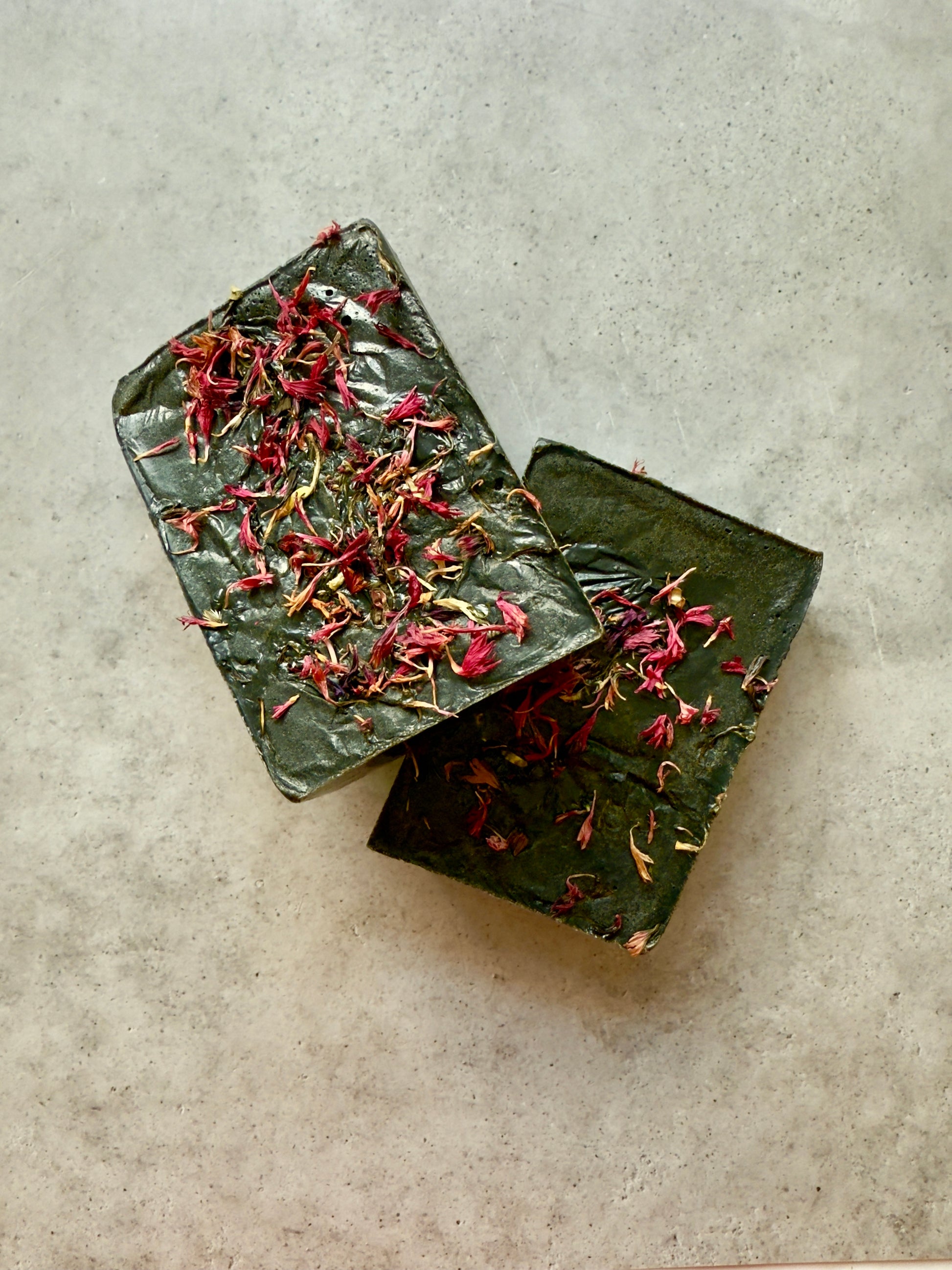 Dark green rectangular soap bar topped with bright pink cornflower petals, shown on a smooth stone-textured background.