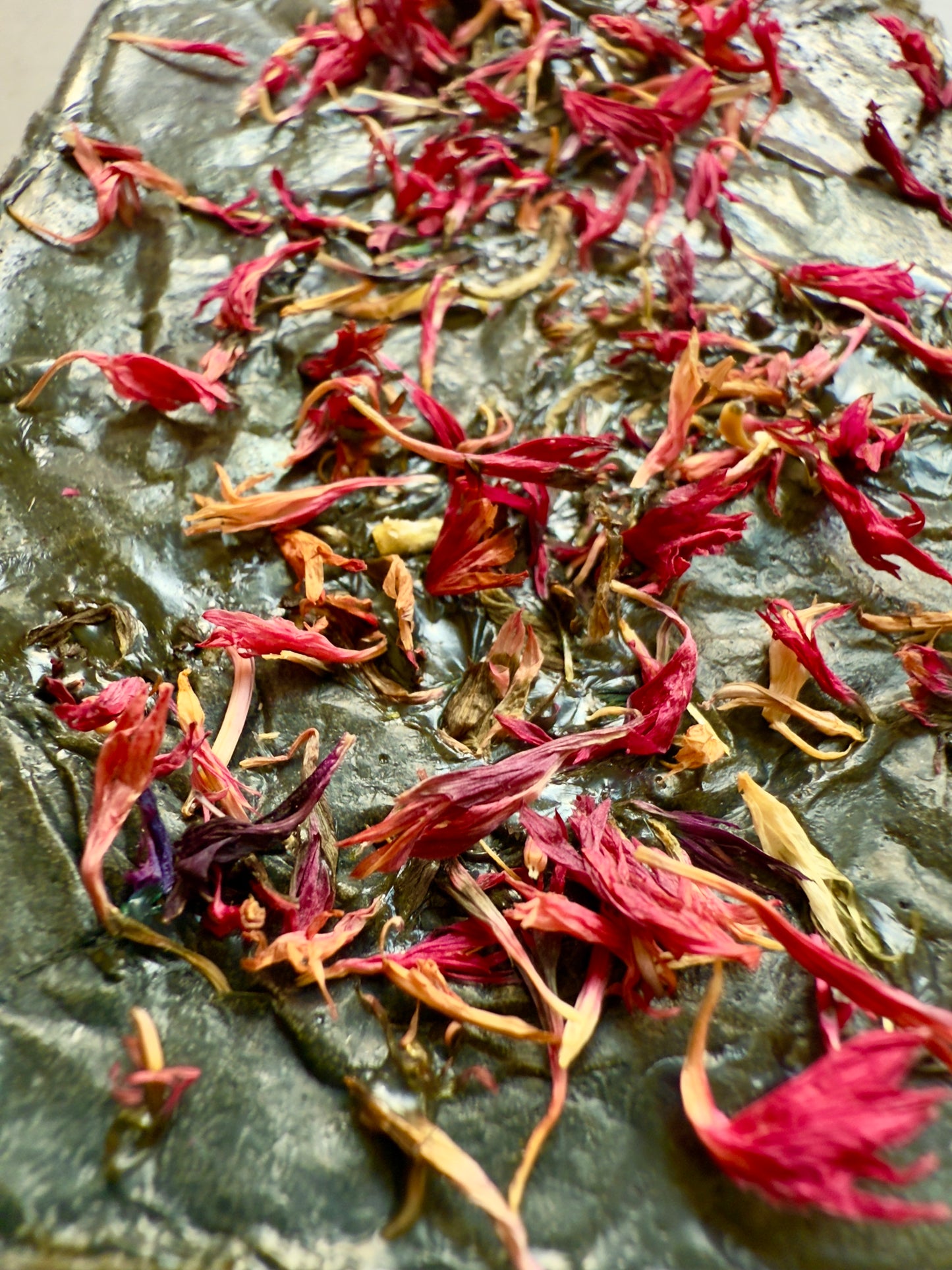 Dark green rectangular soap bar topped with bright pink cornflower petals, shown on a smooth stone-textured background.