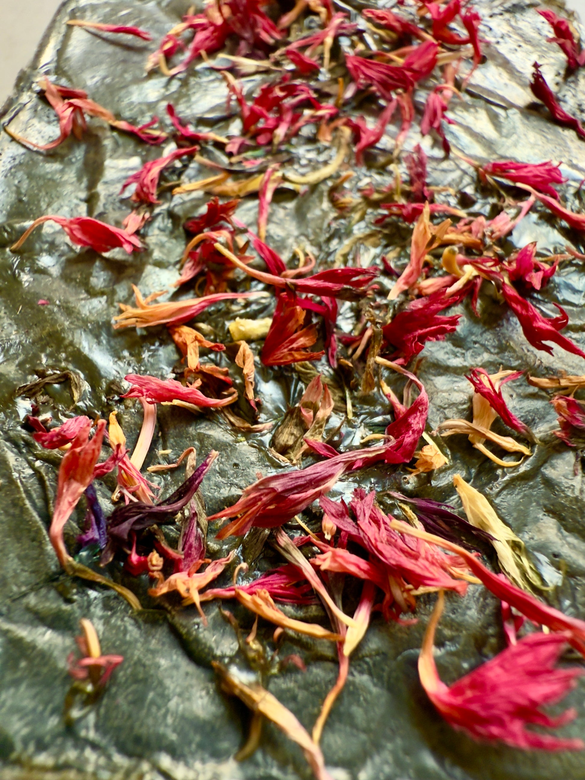 Dark green rectangular soap bar topped with bright pink cornflower petals, shown on a smooth stone-textured background.