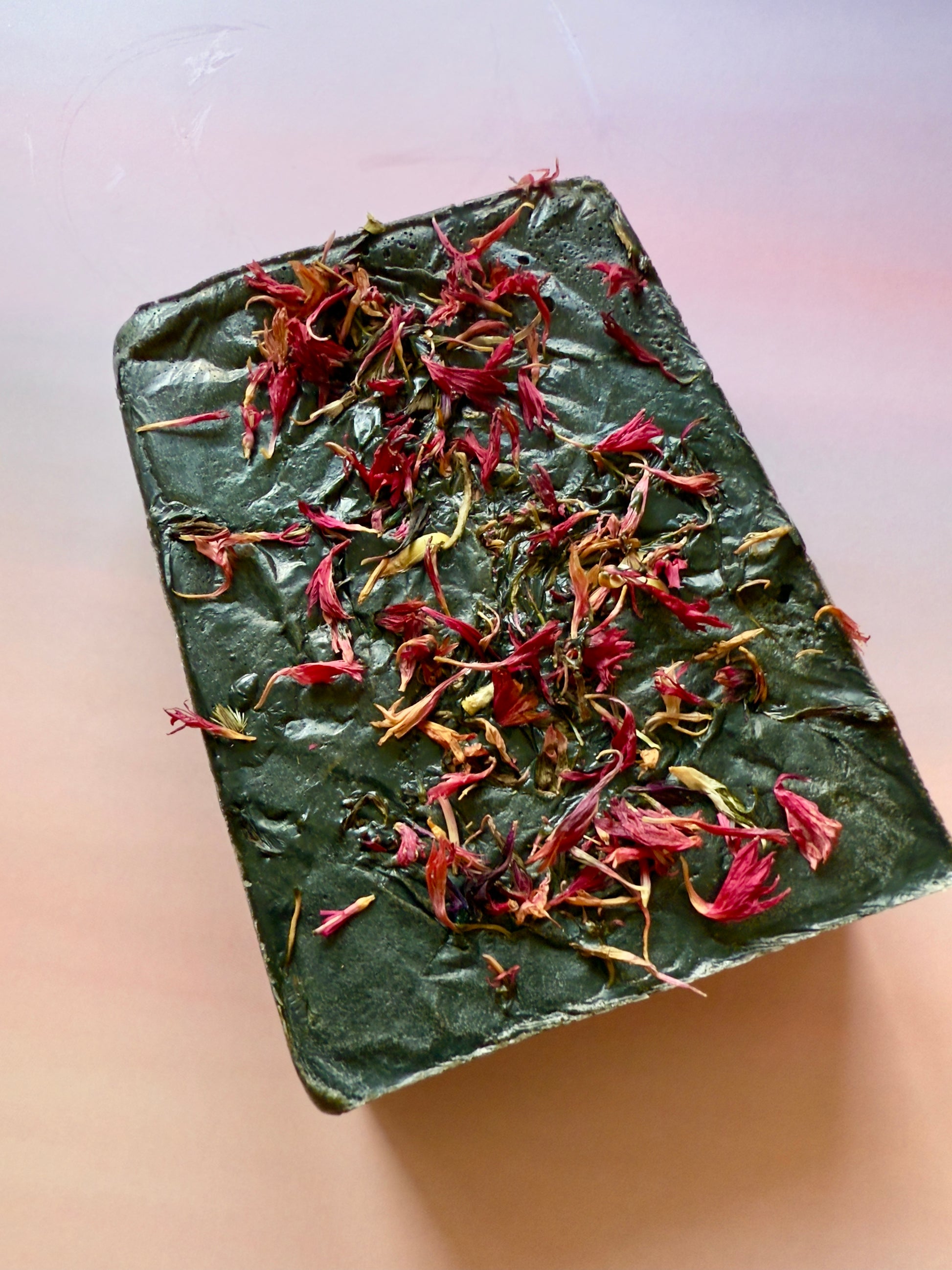 Dark green rectangular soap bar topped with bright pink cornflower petals, shown on a smooth stone-textured background.