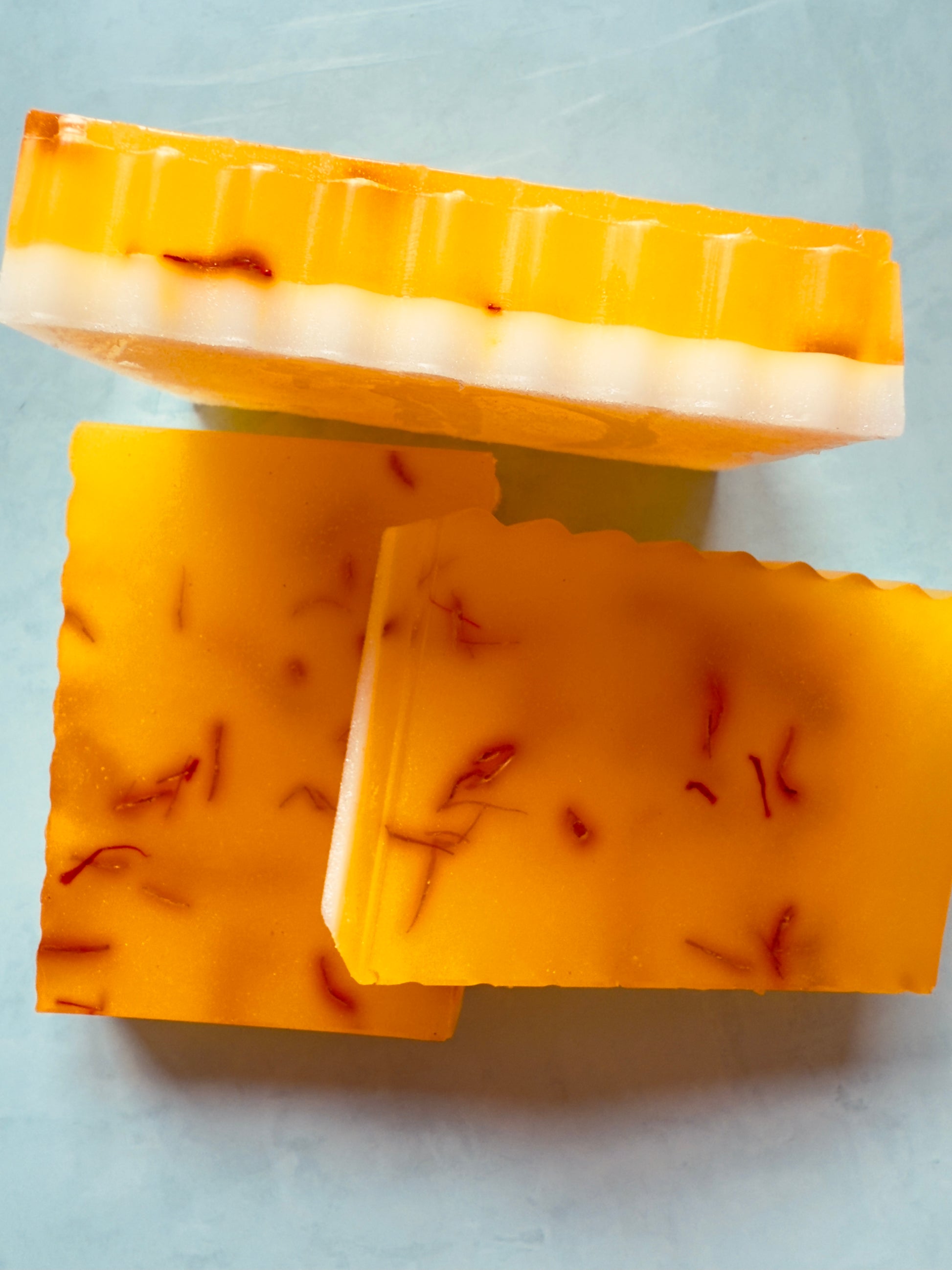 Golden yellow crinkle-cut soap bars with visible saffron threads, layered over a creamy white goat milk base, photographed on a soft gray background.