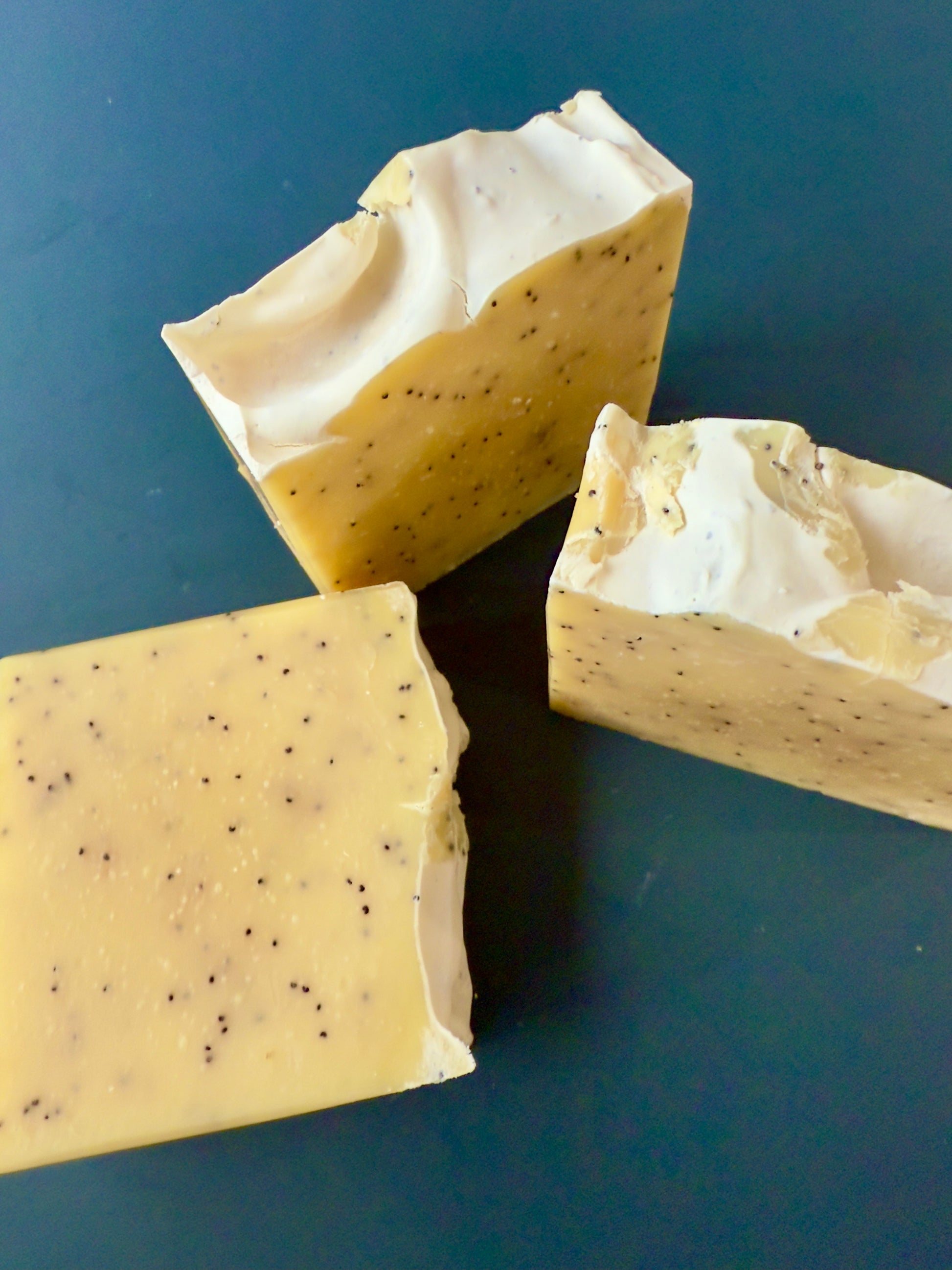 Lemon-colored soap bars with poppy seeds, displayed on a dark teal background.