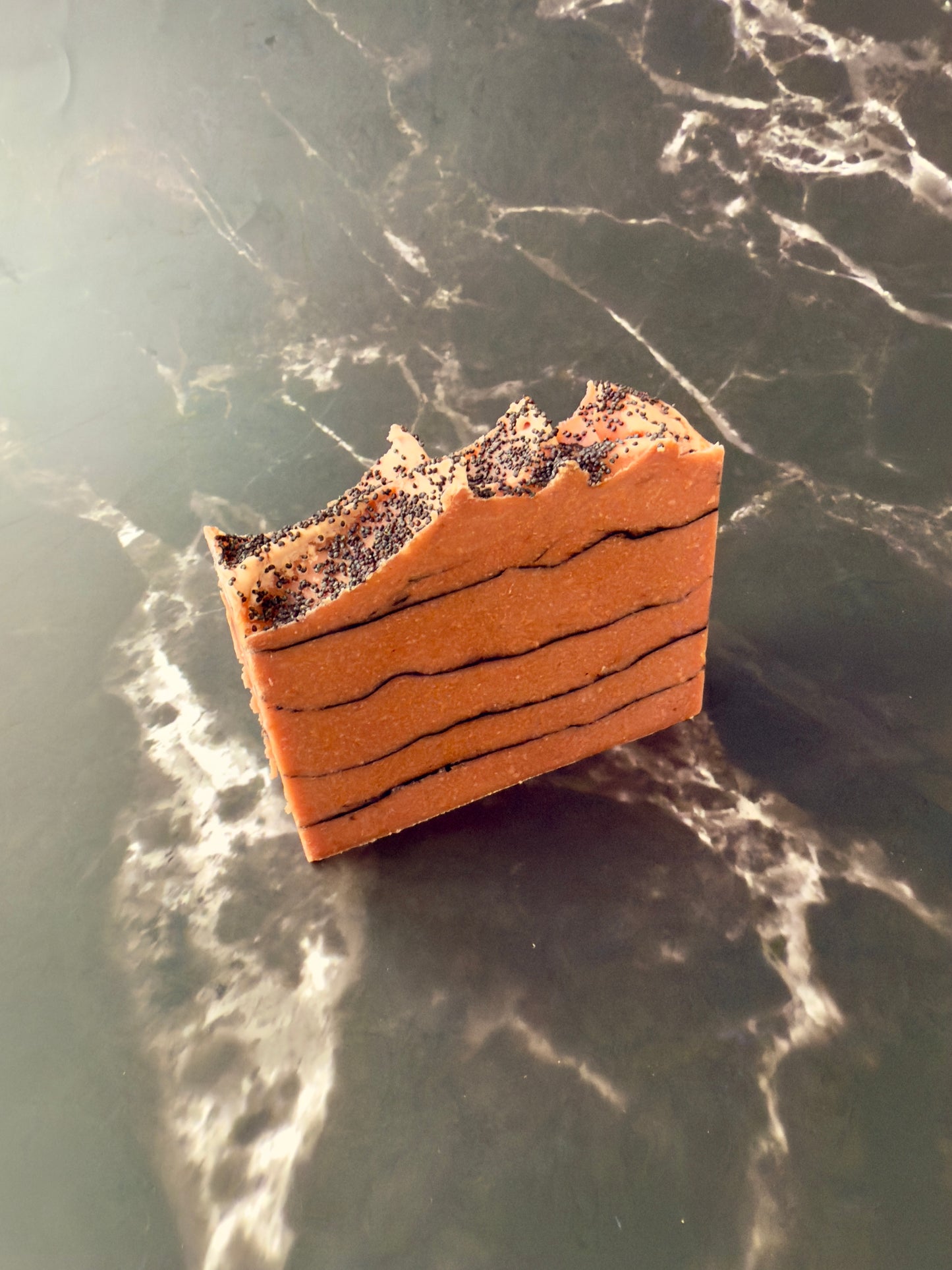 Brick-red Tomato cold process soap with black charcoal pencil lines and poppy-seed top, displayed on dark marble.