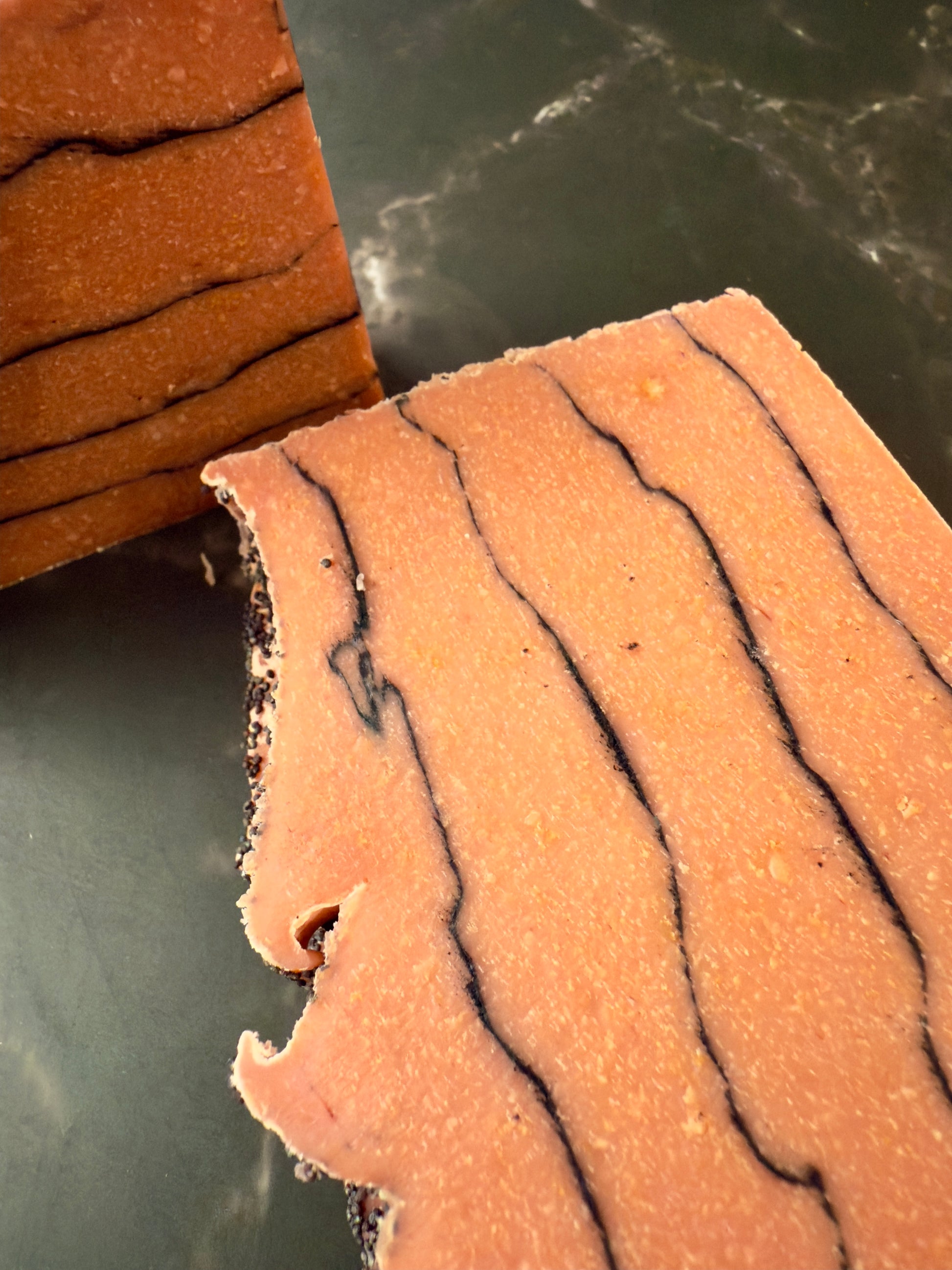 Brick-red Tomato cold process soap with black charcoal pencil lines and poppy-seed top, displayed on dark marble.