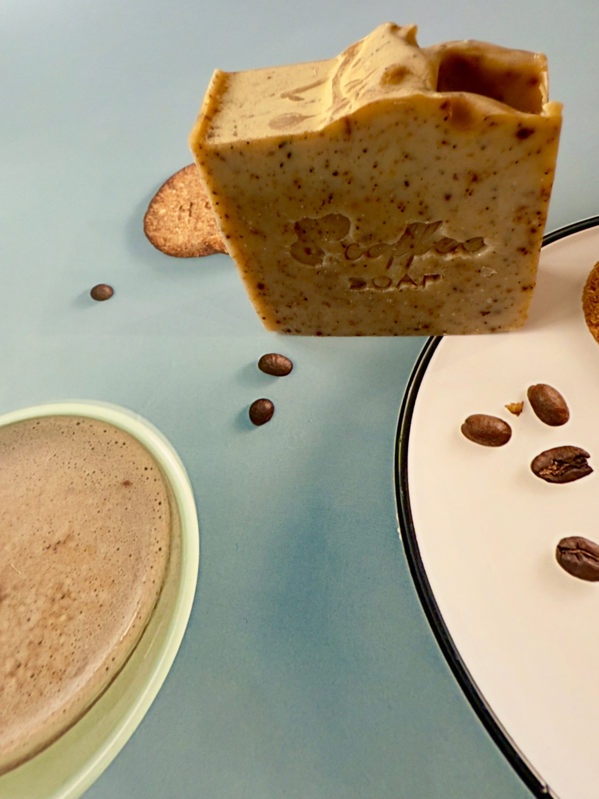 Handcrafted coffee soap bar with dry coffee specks and textured top, photographed with coffee beans and latte elements.