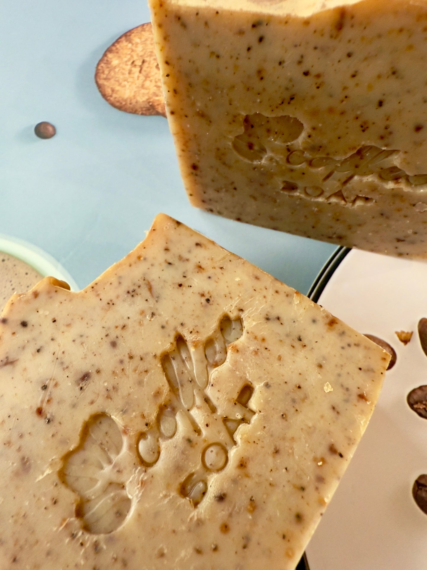 Handcrafted coffee soap bar with dry coffee specks and textured top, photographed with coffee beans and latte elements.