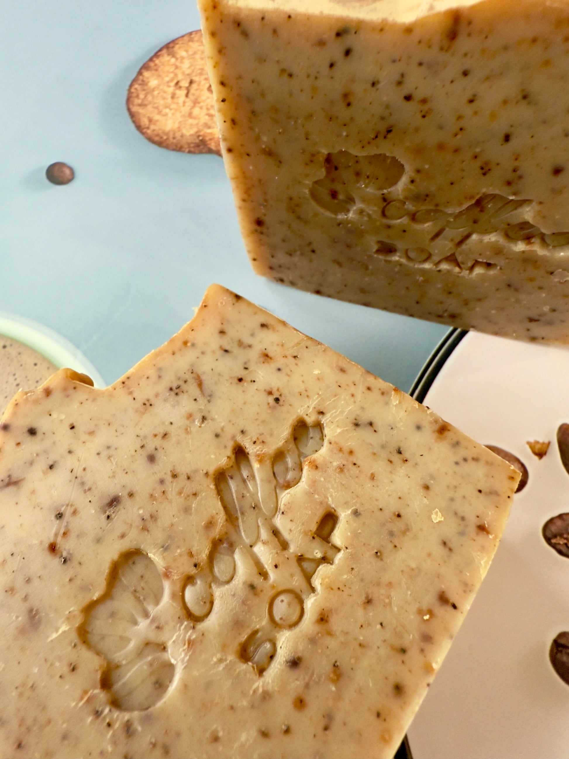 Handcrafted coffee soap bar with dry coffee specks and textured top, photographed with coffee beans and latte elements.