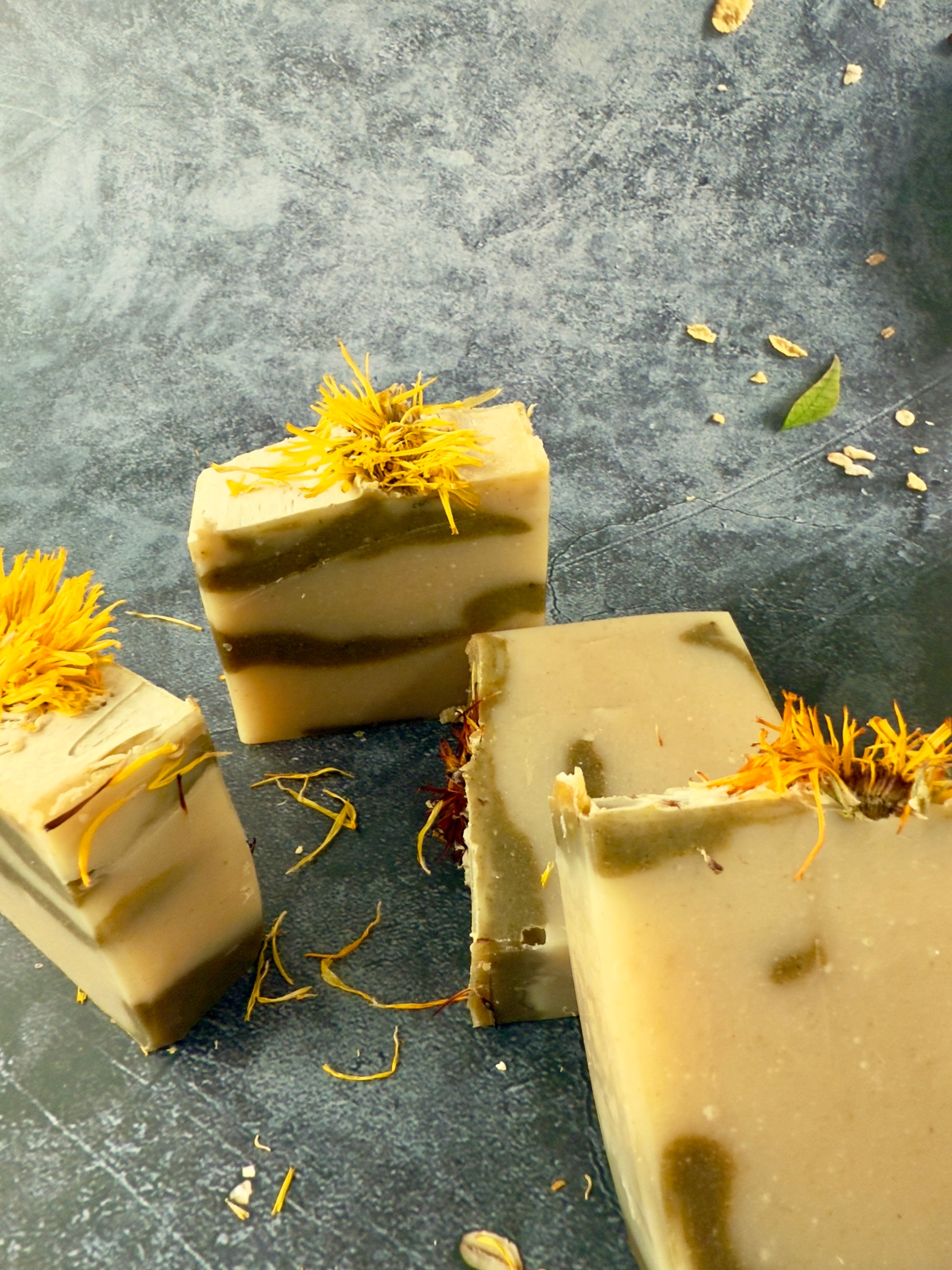 Three bars of soap with yellow flower petals on a gray surface. Layered facial soap bar with calendula petals and natural oatmeal tones.