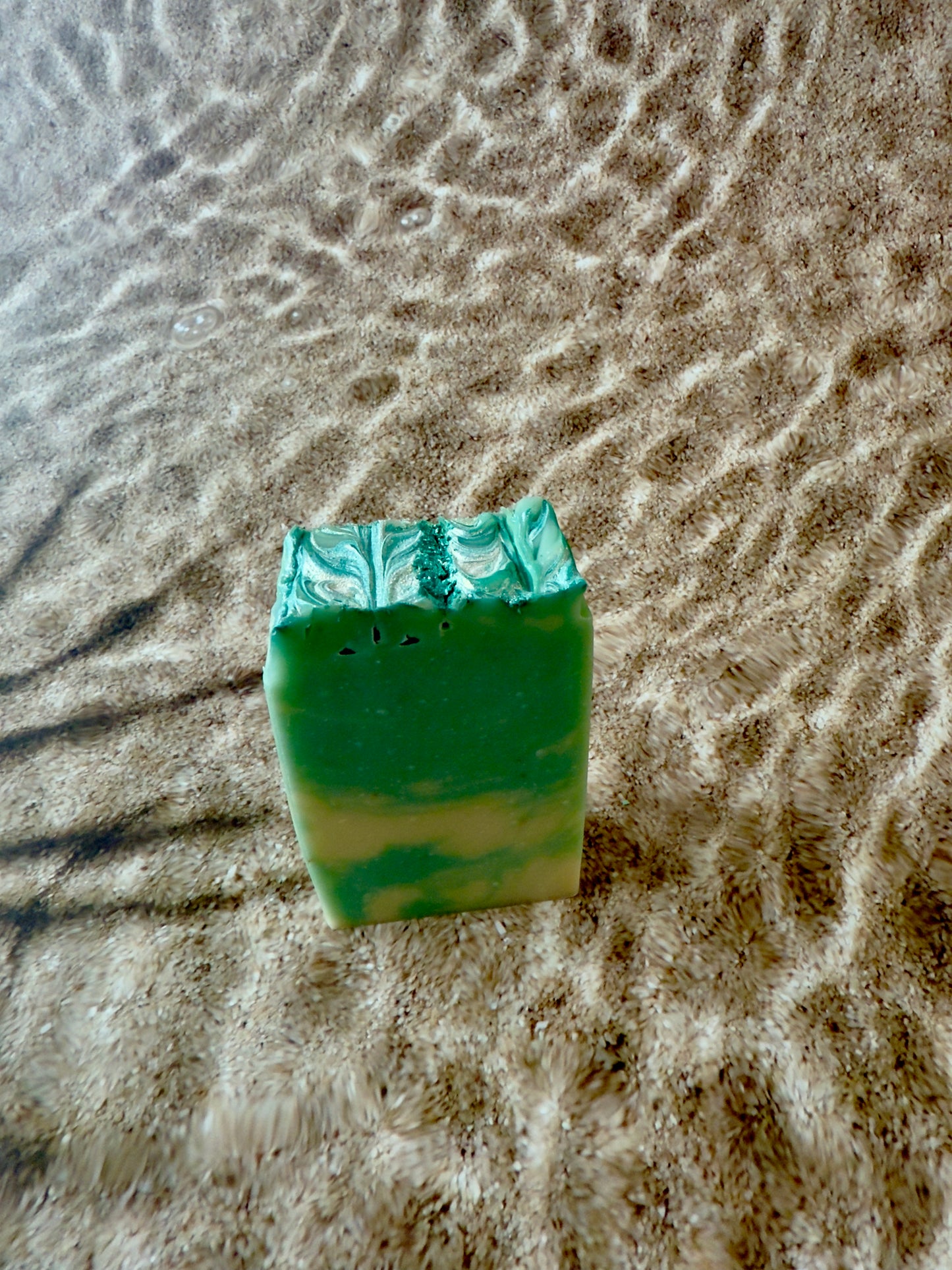 A turquoise and cream swirled Seaside soap bar with a shimmering textured top, photographed on sand.