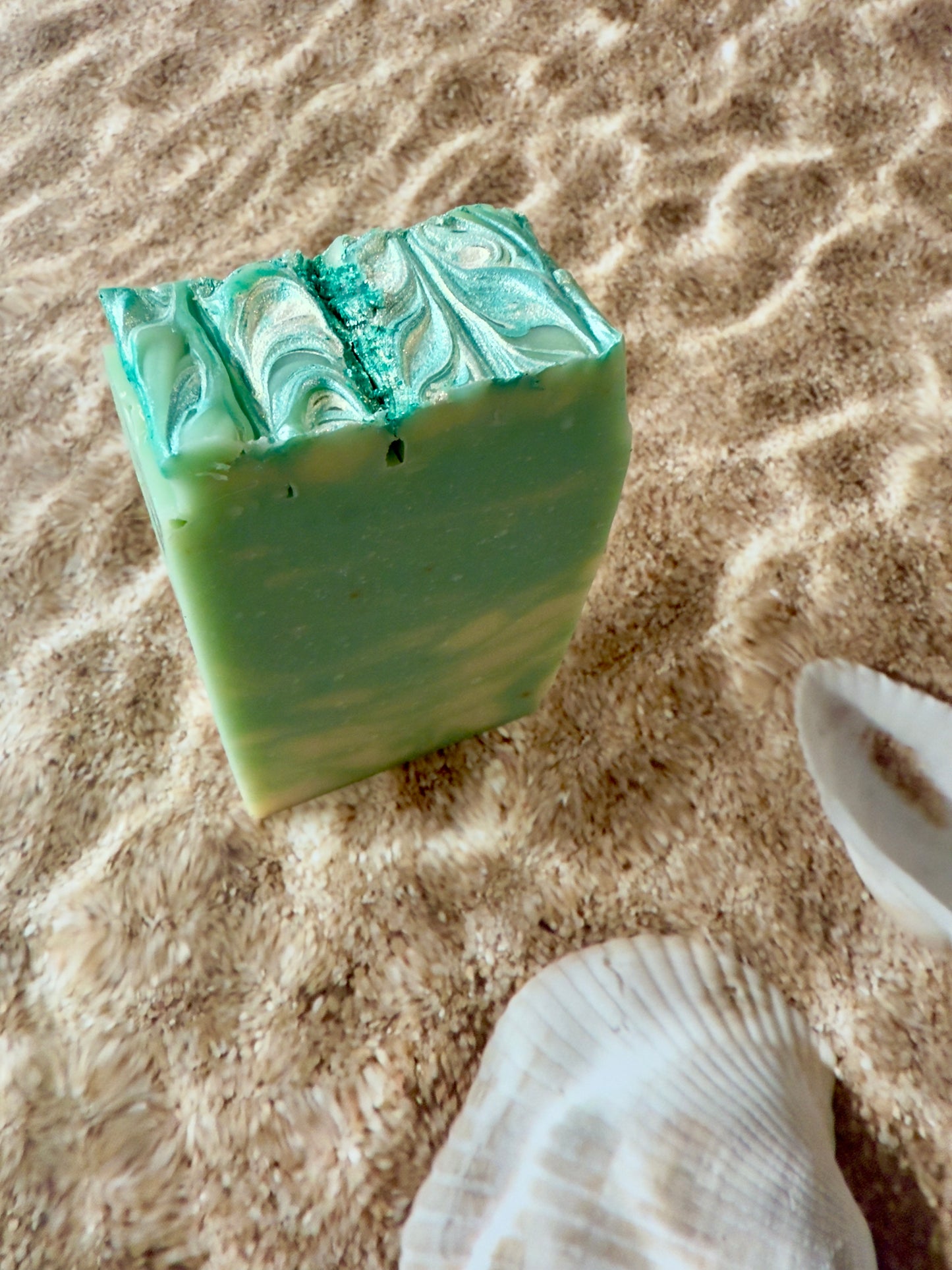 Green soap bar with marbled pattern on sand with a shell nearby