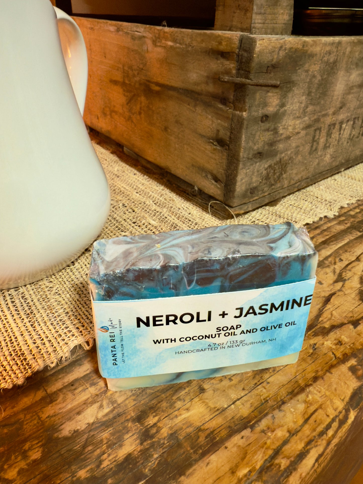 Bar of soap labeled 'Neroli + Jasmine' on a wooden surface with a rustic background. Blue and charcoal swirled Neroli + Jasmine cold process soap bar with label, displayed on rustic wood.