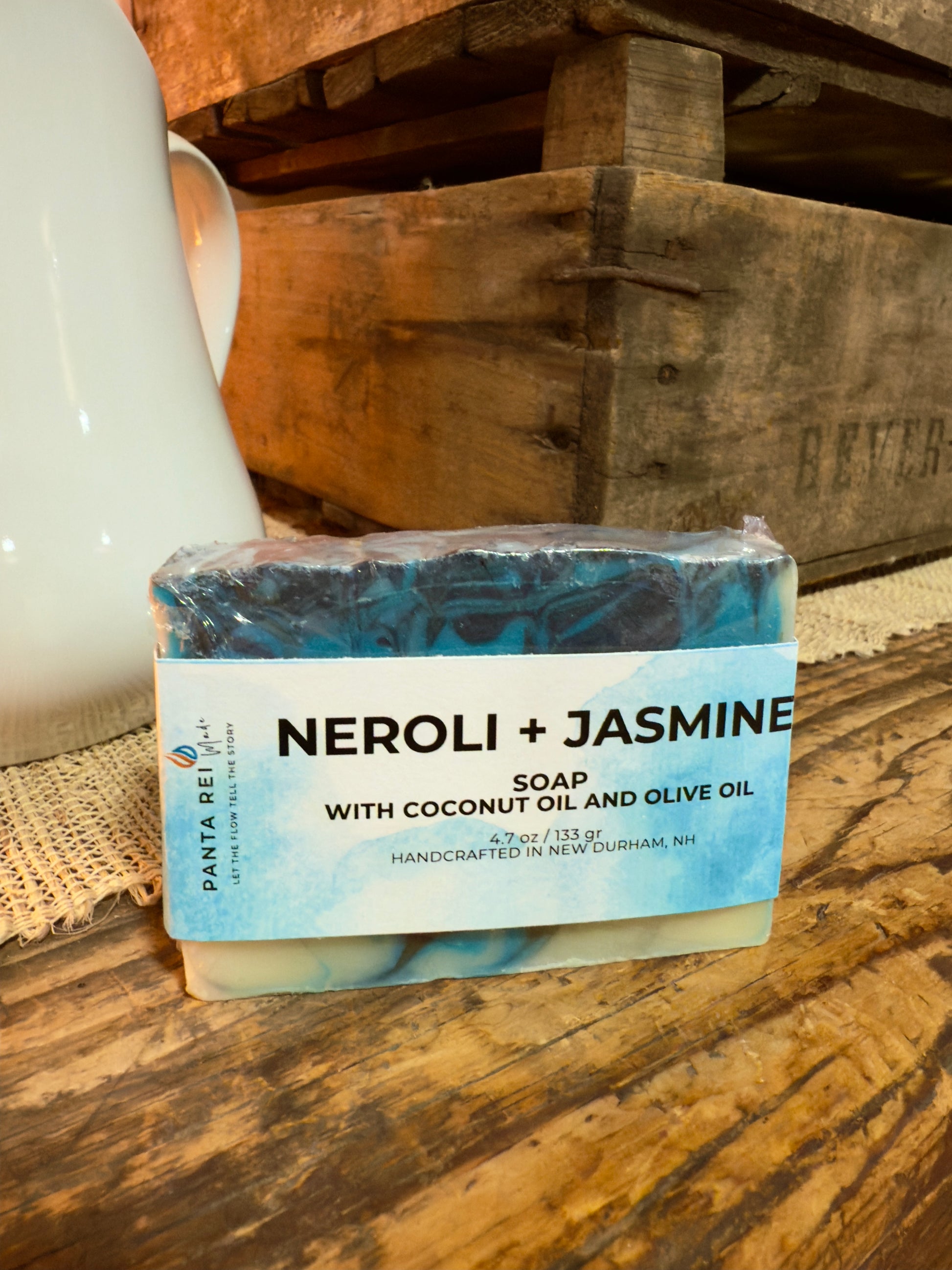 Bar of soap labeled 'Neroli + Jasmine' on a wooden surface with a rustic background. Blue and charcoal swirled Neroli + Jasmine cold process soap bar with label, displayed on rustic wood.