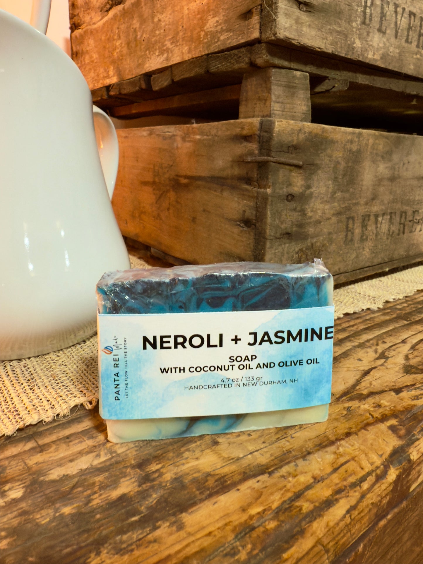 Bar of soap labeled 'Neroli + Jasmine' on a wooden surface with a rustic background. Blue and charcoal swirled Neroli + Jasmine cold process soap bar with label, displayed on rustic wood.
