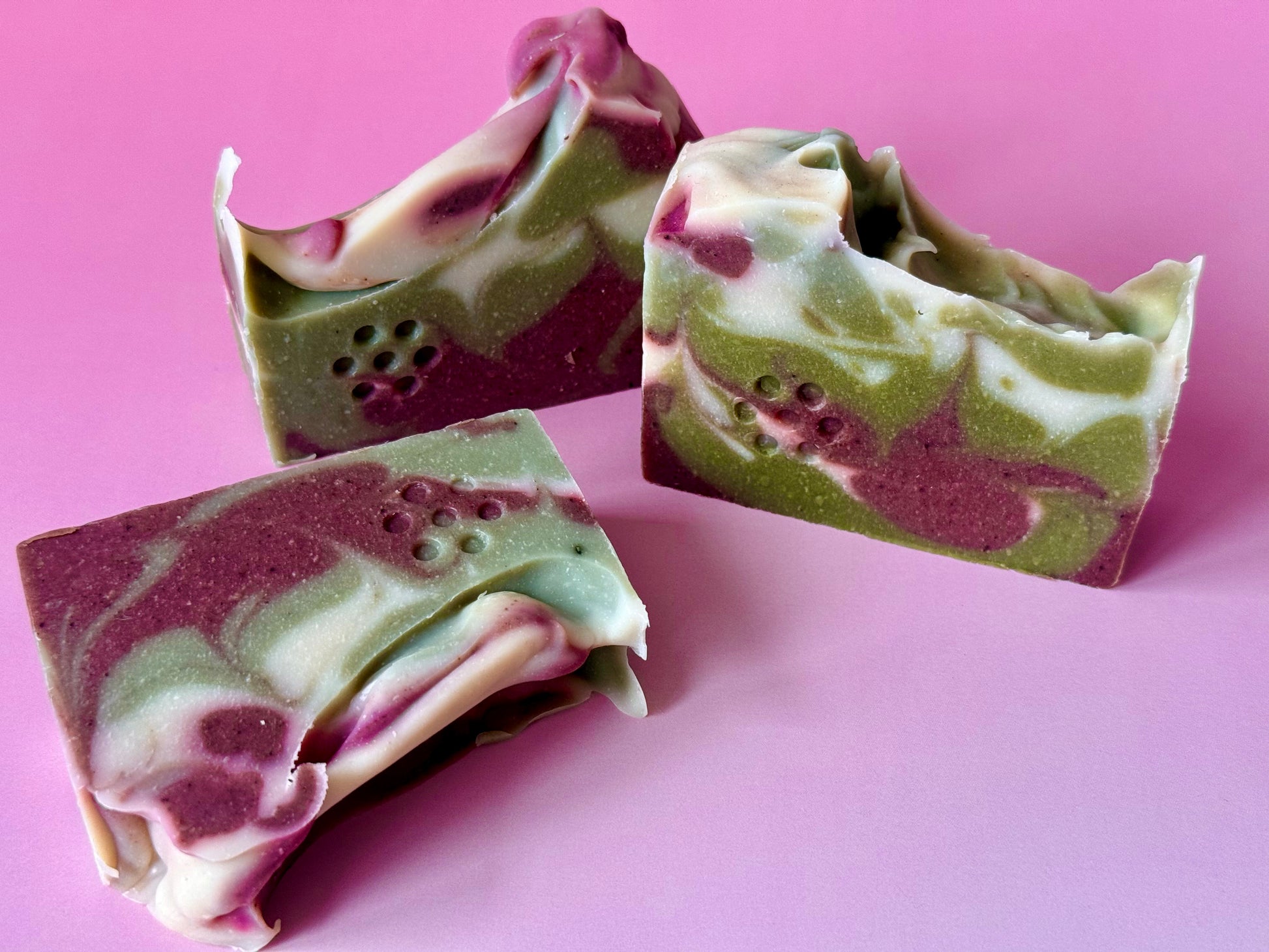 Close-up of green and brown swirled soap bars on a pink background. Swirled green, white, and plum artisan soap bar with a textured top on a pink backdrop.