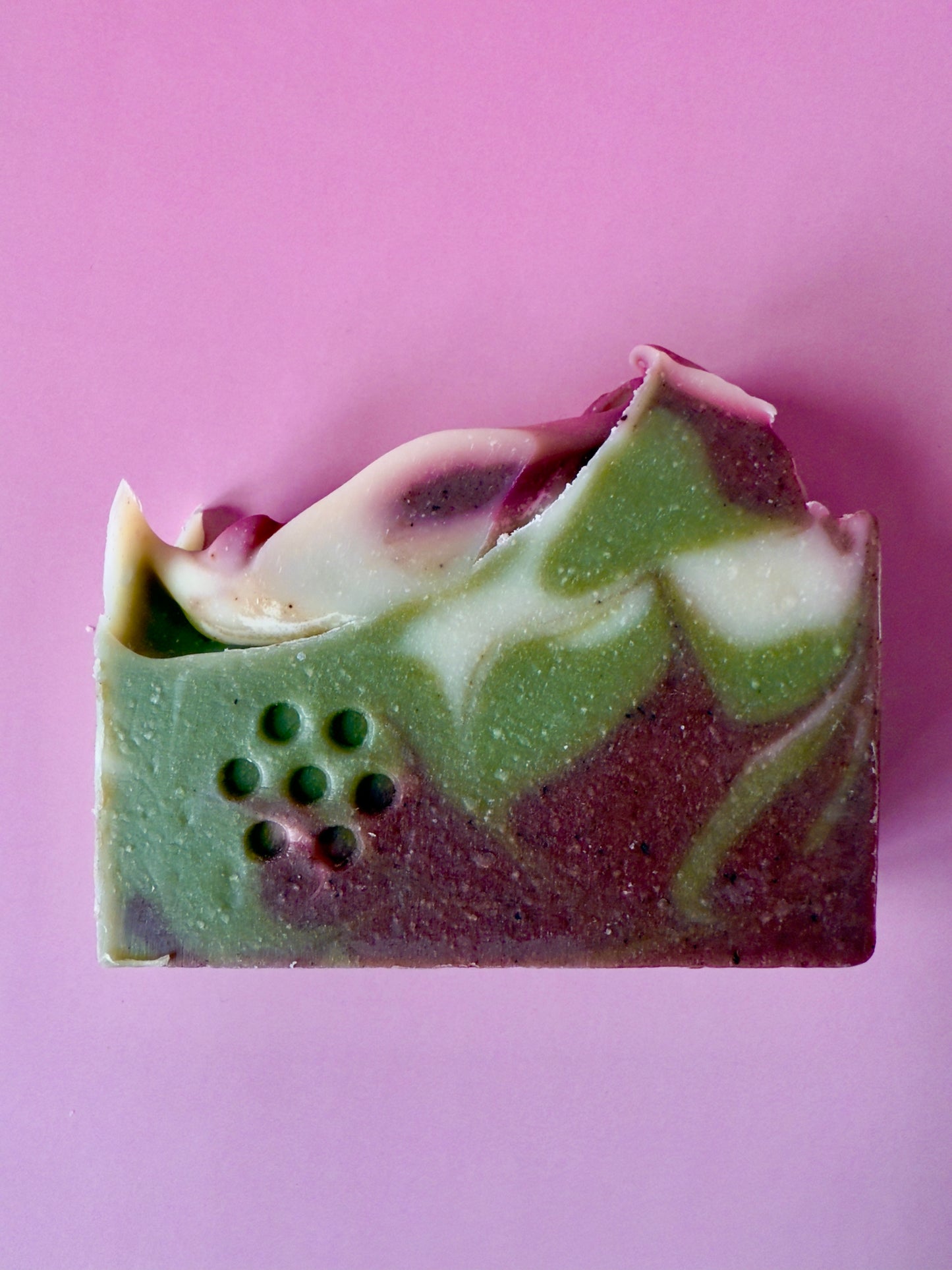 Close-up of green and brown swirled soap bars on a pink background. Swirled green, white, and plum artisan soap bar with a textured top on a pink backdrop.