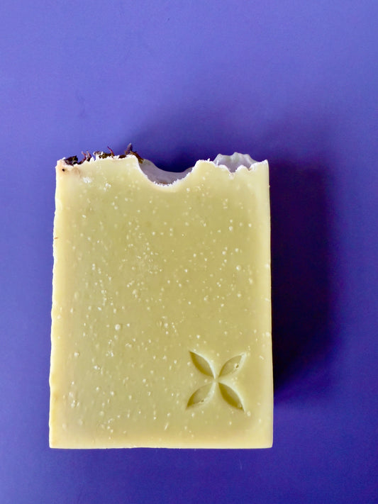 Green artisan soap bar with lavender buds on top and a carved floral stamp on a purple background.