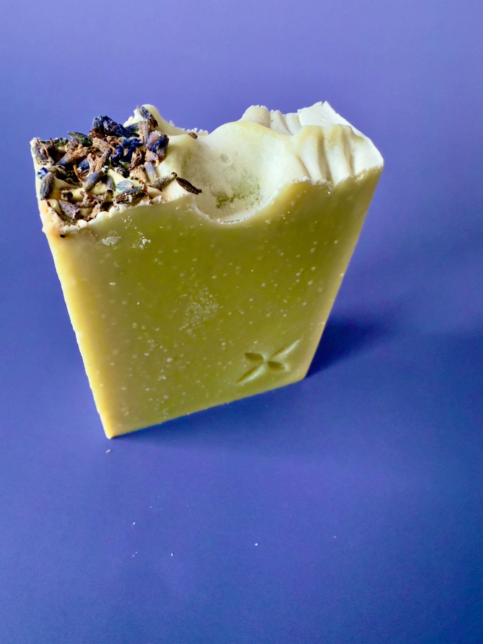 Green artisan soap bar with lavender buds on top and a carved floral stamp on a purple background.
