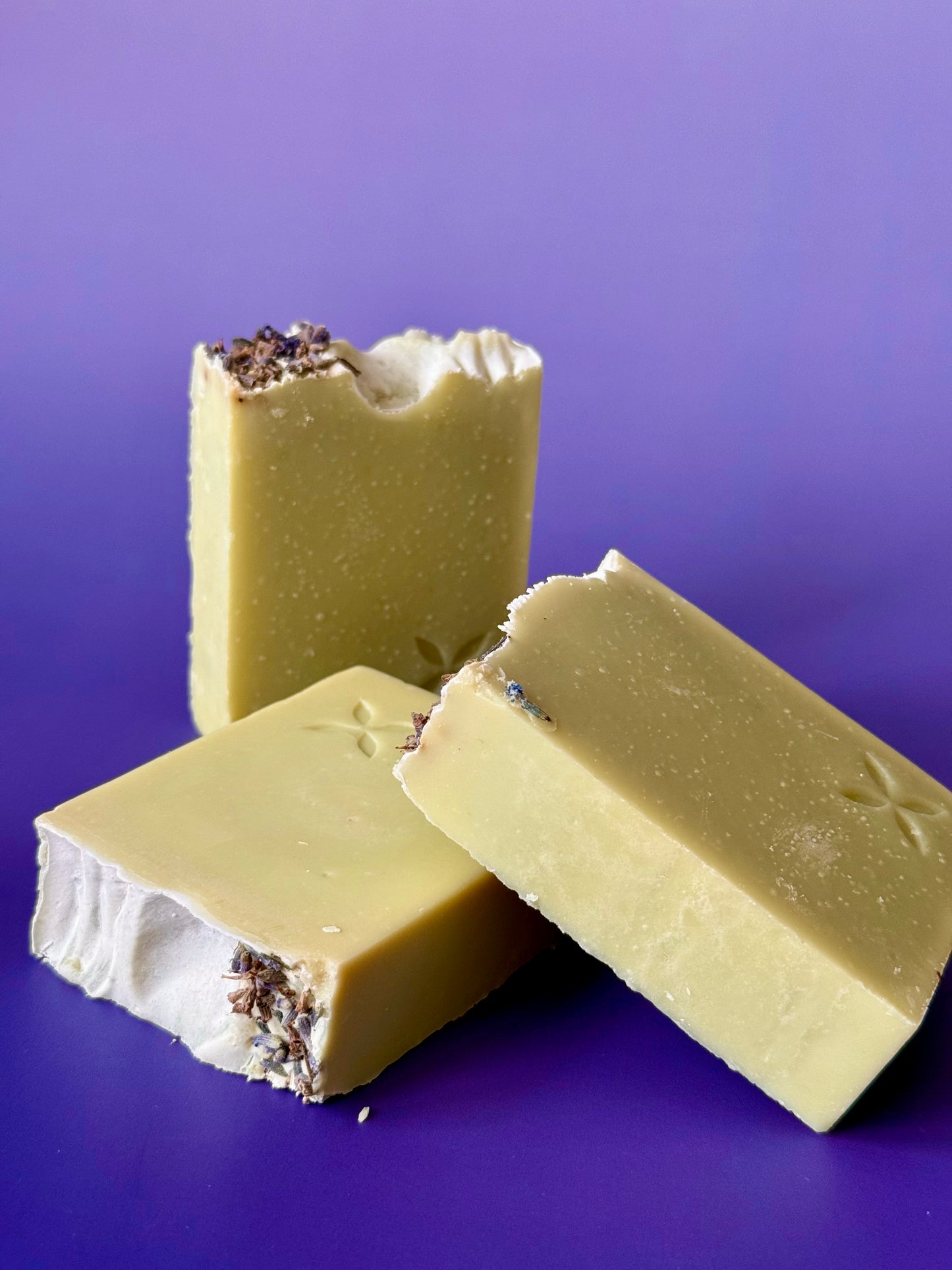 Green artisan soap bar with lavender buds on top and a carved floral stamp on a purple background.