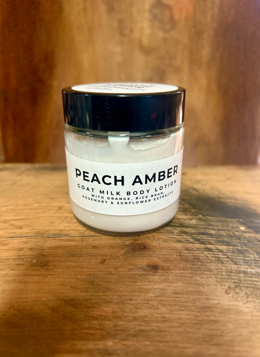 Peach Amber goat milk body lotion in a clear jar with a black lid and minimalist label, photographed on a warm wood background.
