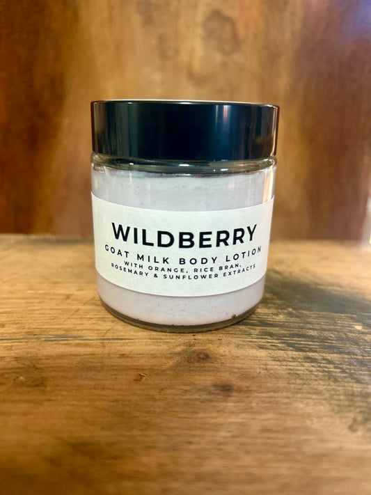 Wildberry goat milk body lotion in a clear jar with a black lid and minimalist label, photographed on a warm wood background.