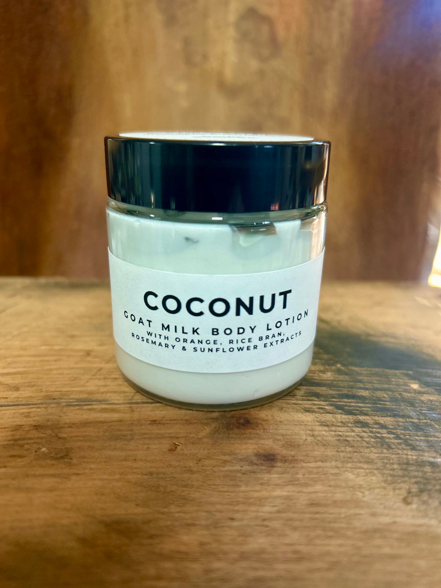 Coconut goat milk body lotion in a clear jar with a black lid and minimalist label, photographed on a warm wooden surface.
