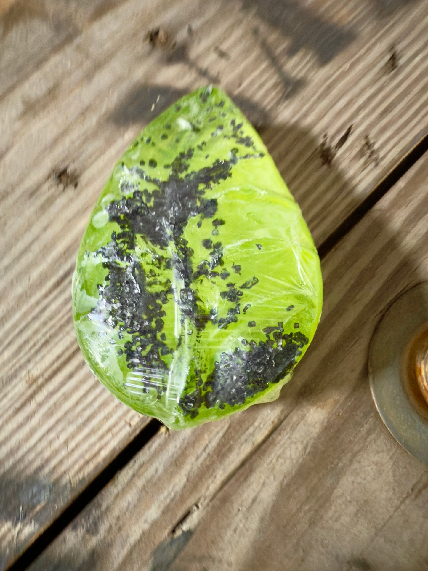 Bright green leaf-shaped soap wrapped in clear film, topped with coarse black salt, photographed on rustic wooden boards.