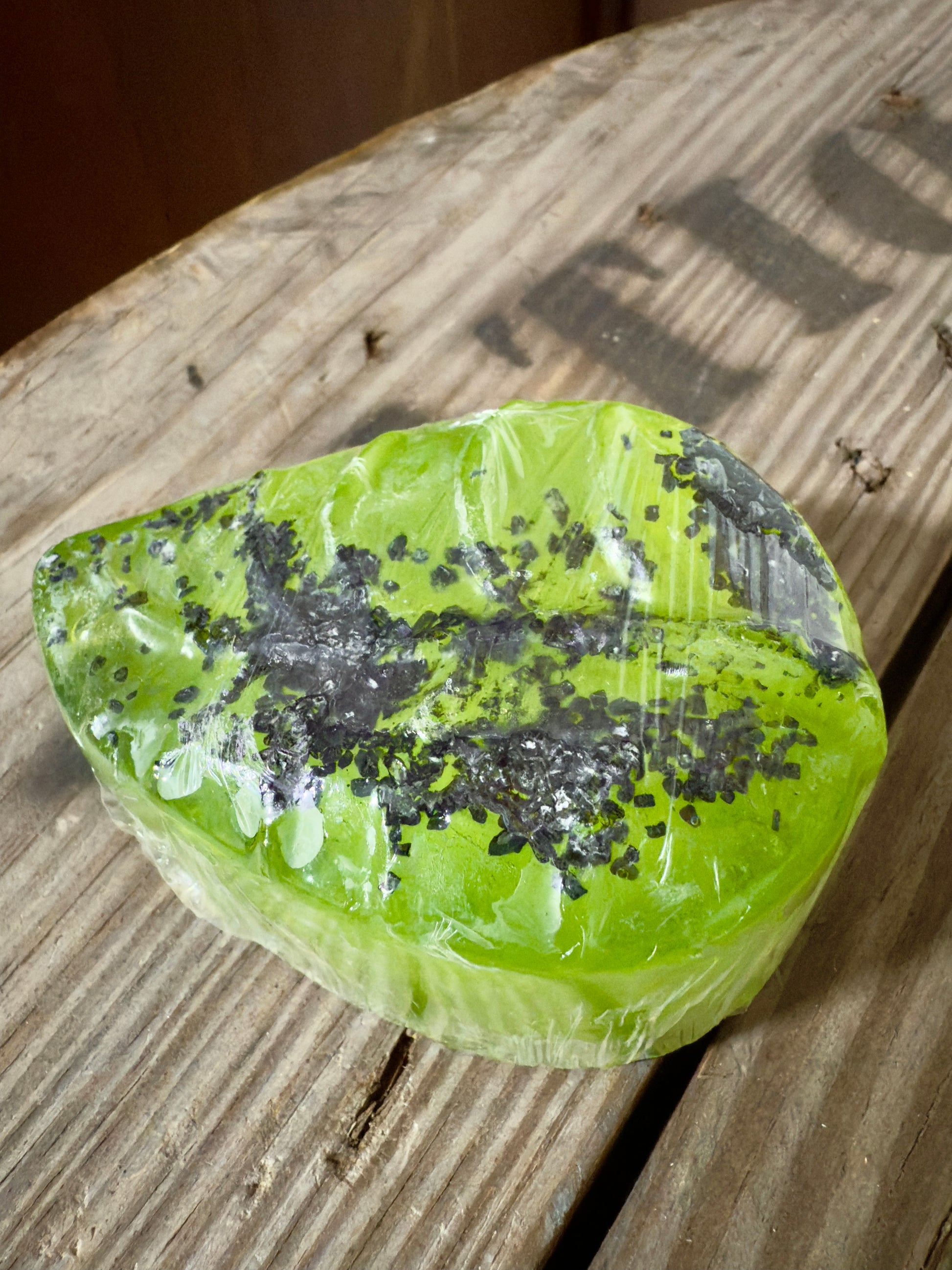Bright green leaf-shaped soap wrapped in clear film, topped with coarse black salt, photographed on rustic wooden boards.