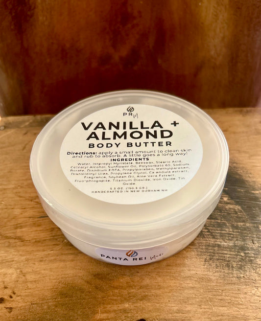 Vanilla + Almond body butter in a clear jar with a white lid and minimalist label, photographed on a warm wood background.