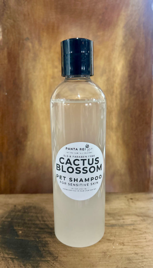 Clear bottle of Cactus Blossom Pet Shampoo with black cap, labeled for sensitive skin, shown on a wooden surface.